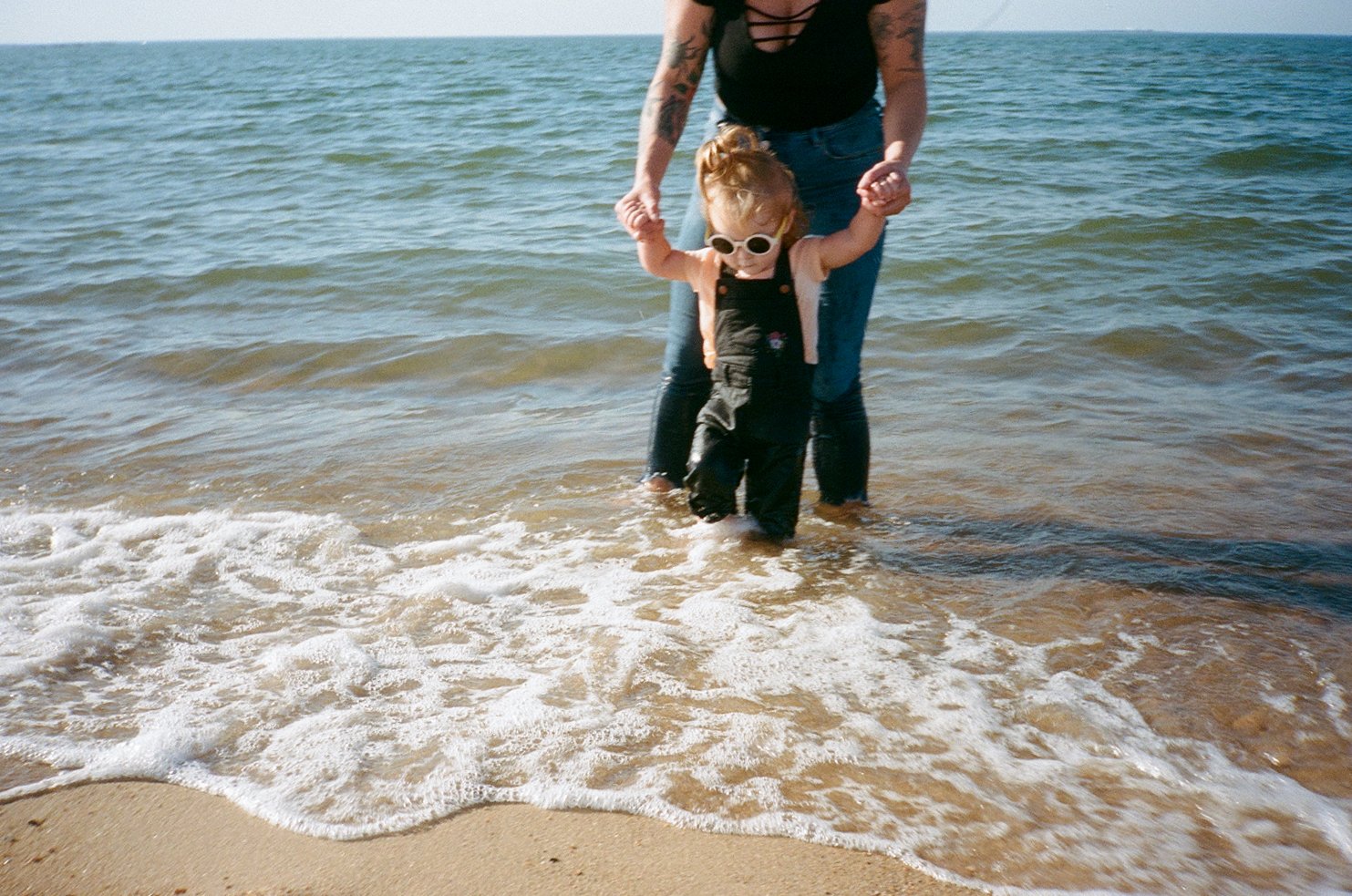 Toddler in sunglasses with romper, held by tattooed adult standing in ocean water at beach.