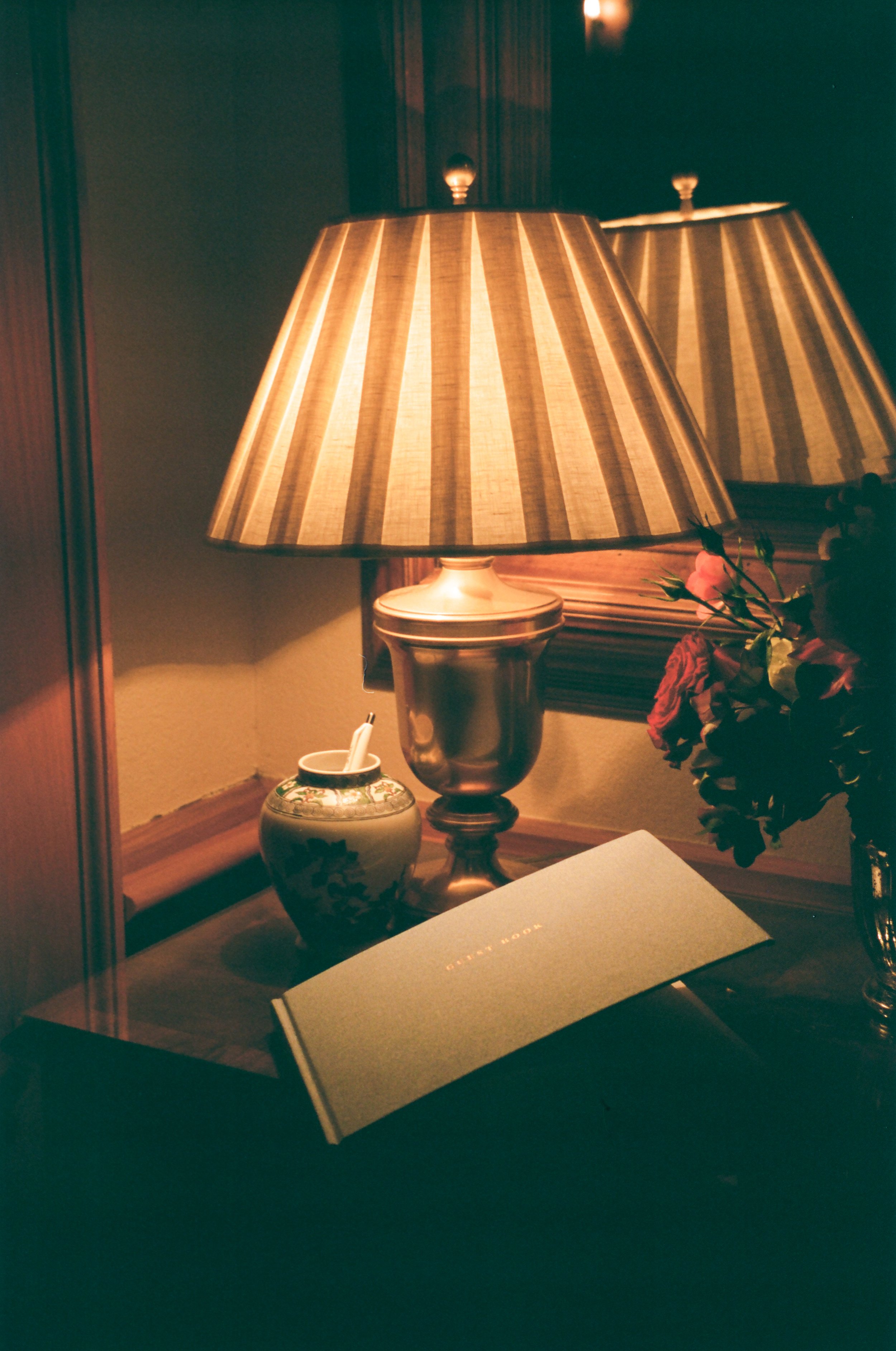 A table lamp with a striped lampshade, a small decorative vase with pens, a bouquet of flowers in a vase, and a green hardcover notebook, all reflected in a mirror on a wooden wall.