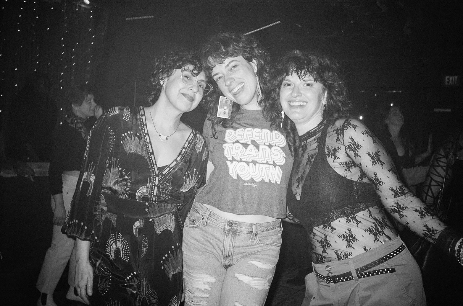 Three smiling people posing together in a black and white photo at a party or gathering. Center person wearing a 'Defend Trans Youth' t-shirt.