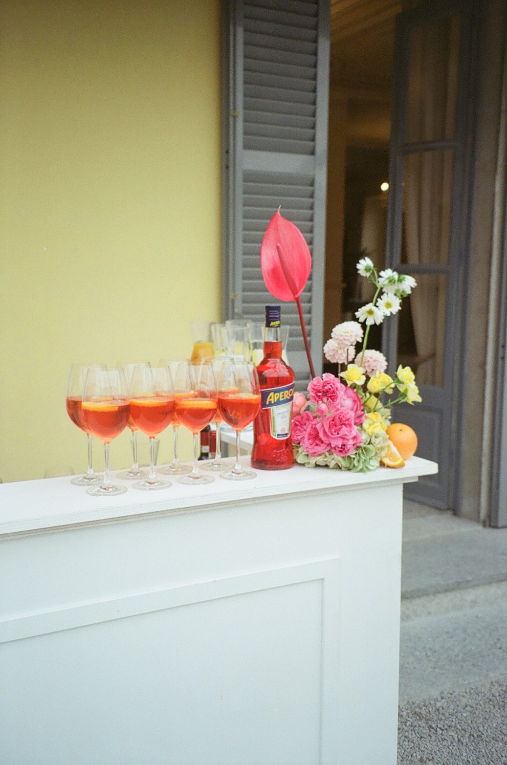 A white bar counter with six glasses of pink-colored drink, a bottle of Aperol, and a floral arrangement including pink, white, and yellow flowers, an orange, and a red decorative leaf, positioned outdoors near a building with closed shutters.