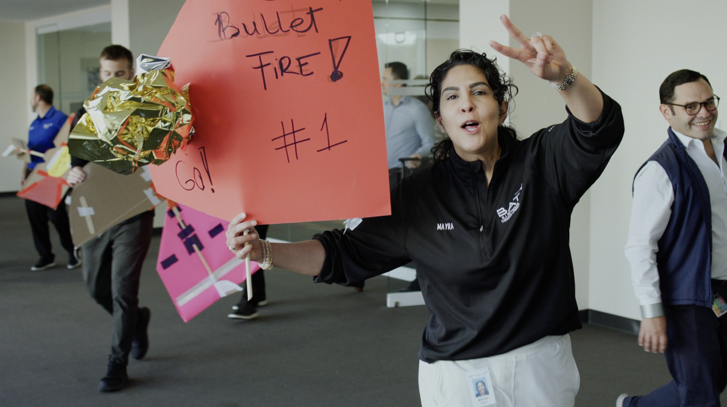 A woman holding a large red sign that reads 'Bullet Fire! #1 GO!' and an wrapped item, while making a peace sign with her fingers. Other individuals in the background are carrying handmade signs and appear to be participating in a protest or rally.