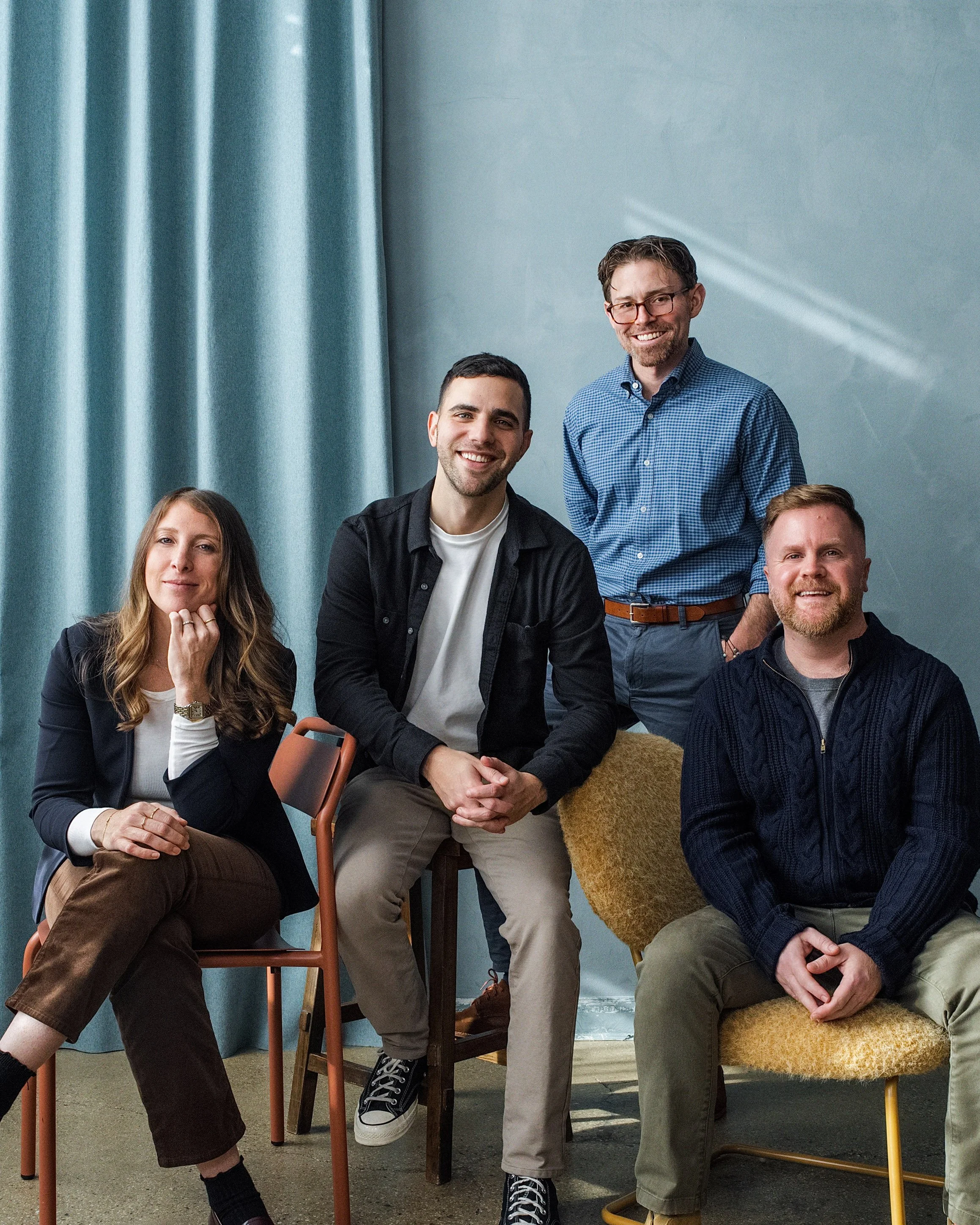 Group of five diverse adults posing together indoors in front of a light blue wall and curtain, smiling and dressed casually.