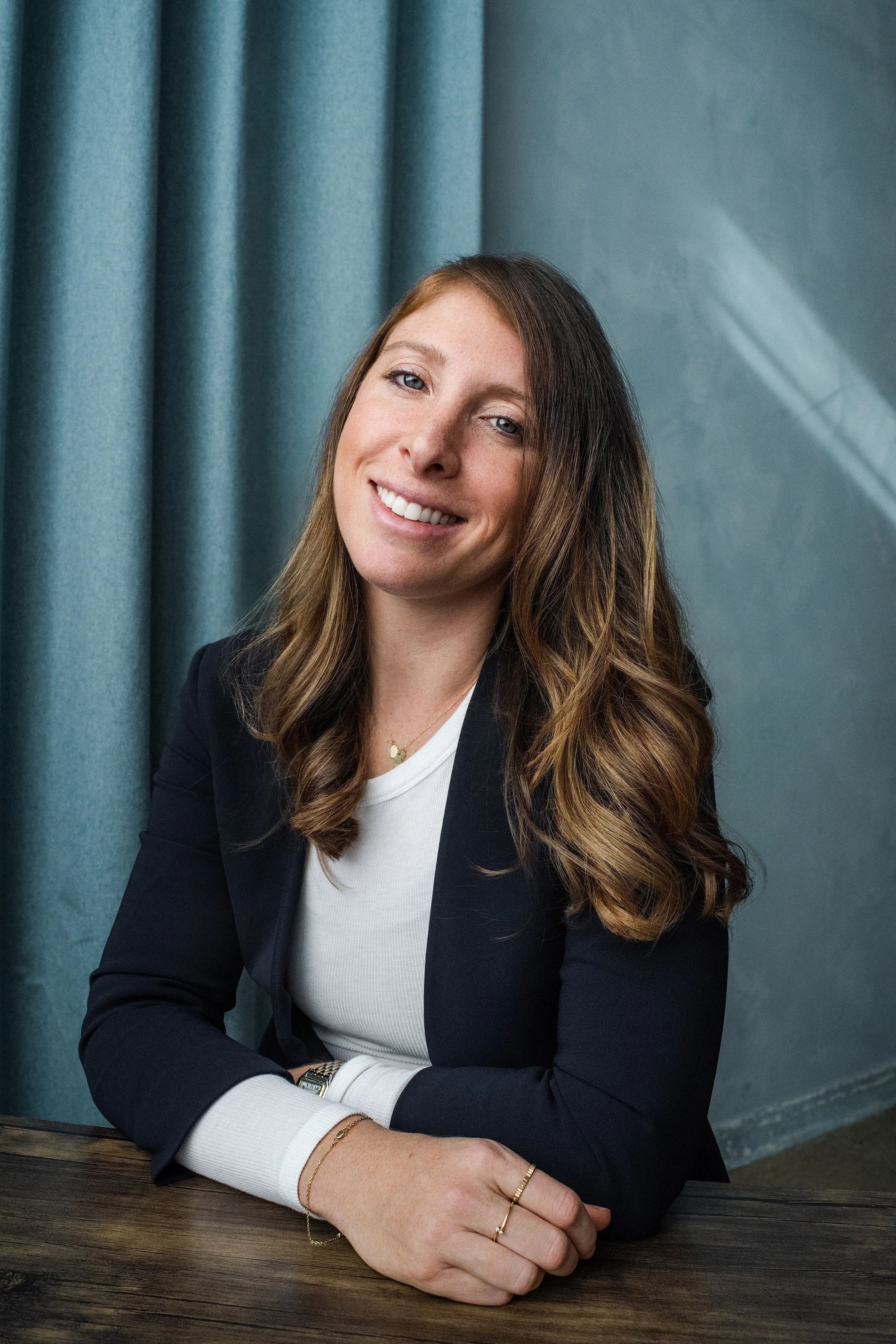 A woman with long, wavy brown hair, smiling, wearing a black blazer and a white shirt, seated at a dark wooden table with teal curtains in the background.