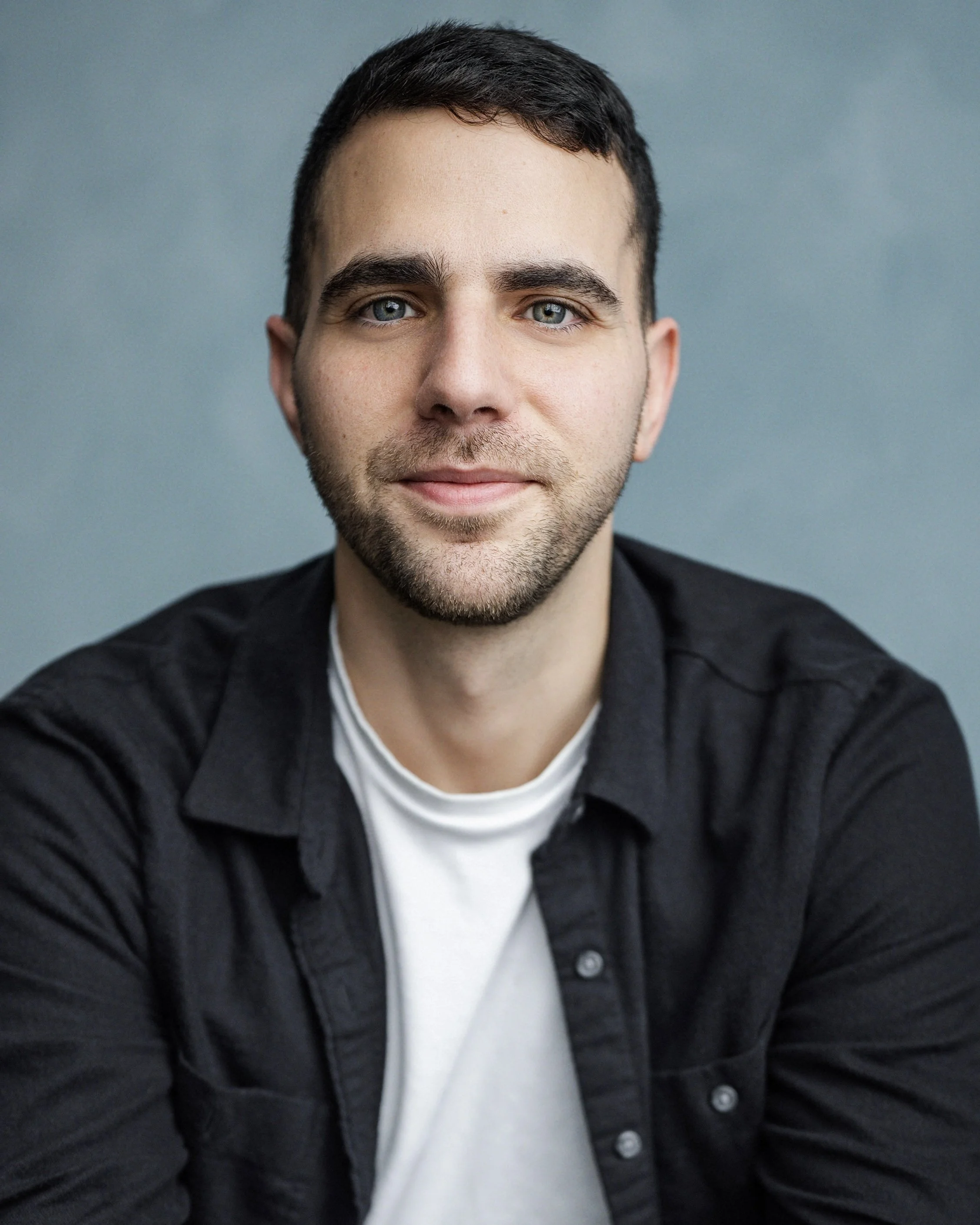 A young man with short dark hair and a beard, sitting at a wooden table, wearing a white t-shirt and a black open shirt, with a neutral blue-gray background.