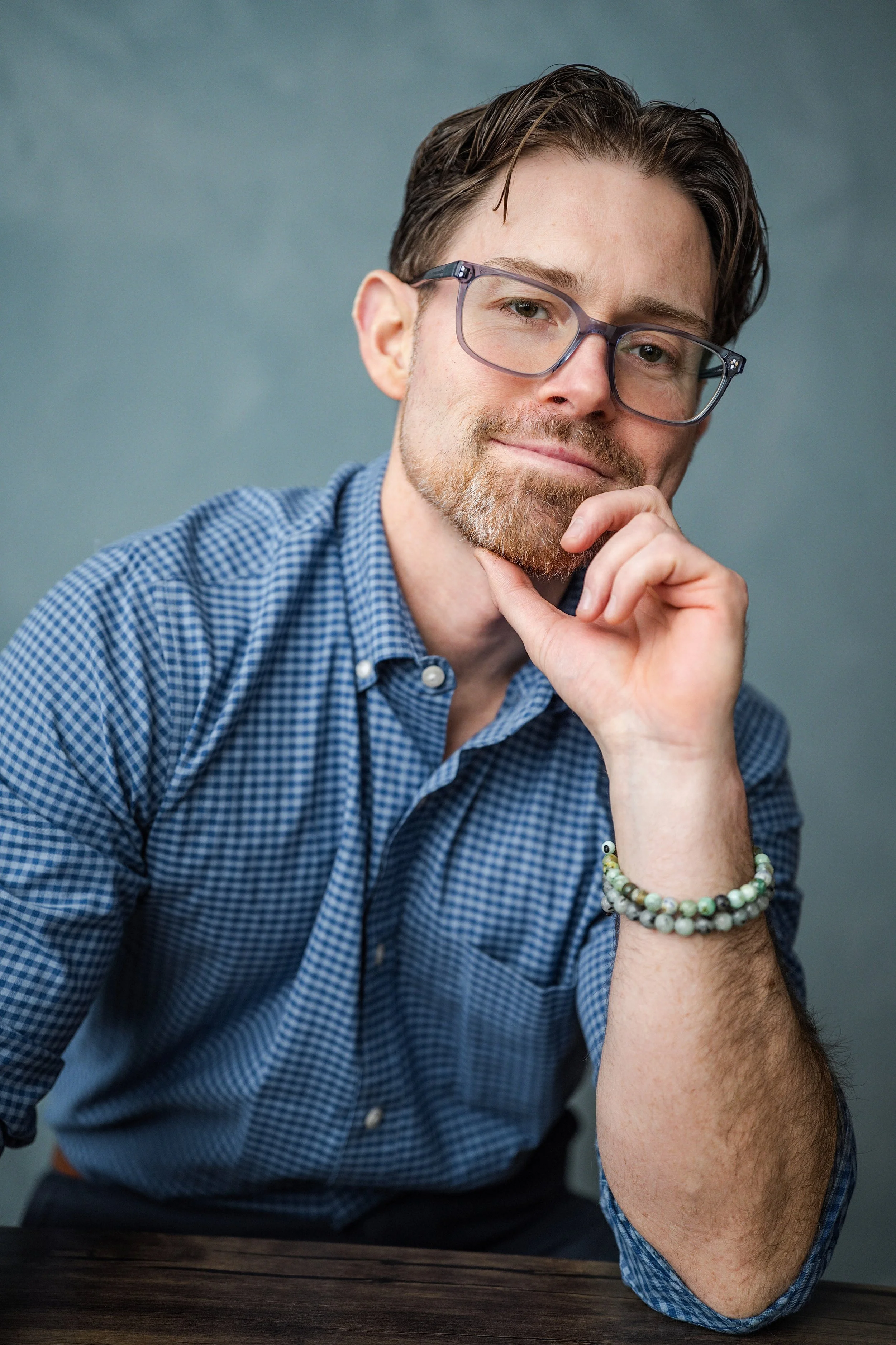 A man with glasses, a beard, and a mustache resting his chin on his hand wearing a blue checkered shirt and bracelets, looking at the camera.