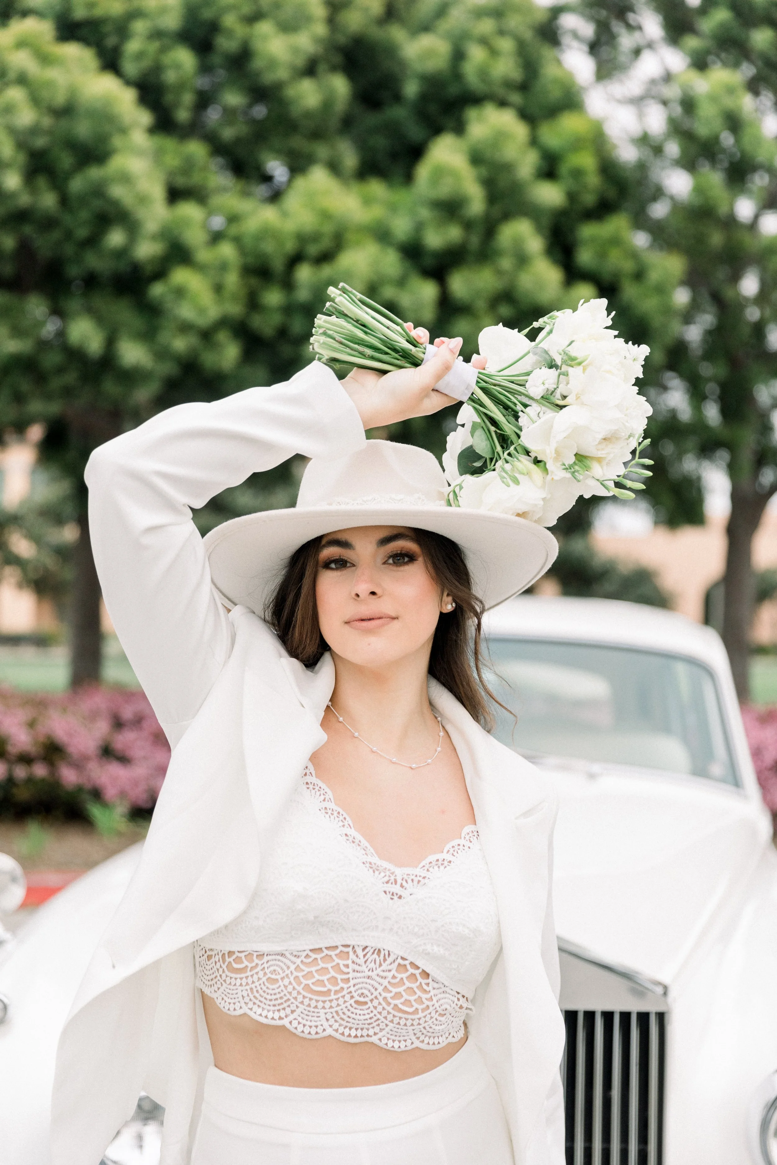 A young woman in a white lace crop top, white jacket, and white wide-brimmed hat holding a bouquet of white flowers above her head outdoors near a vintage white car, with green trees and pink flowers in the background.