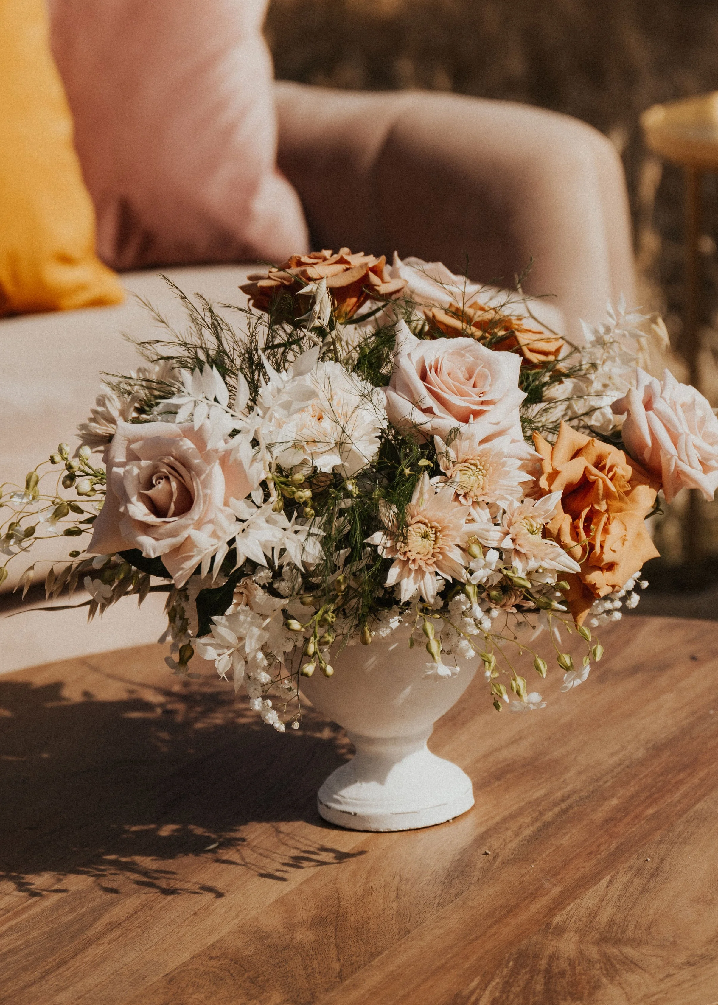 A bouquet of peach and pink roses, white flowers, and greenery in a white vase on a wooden table.