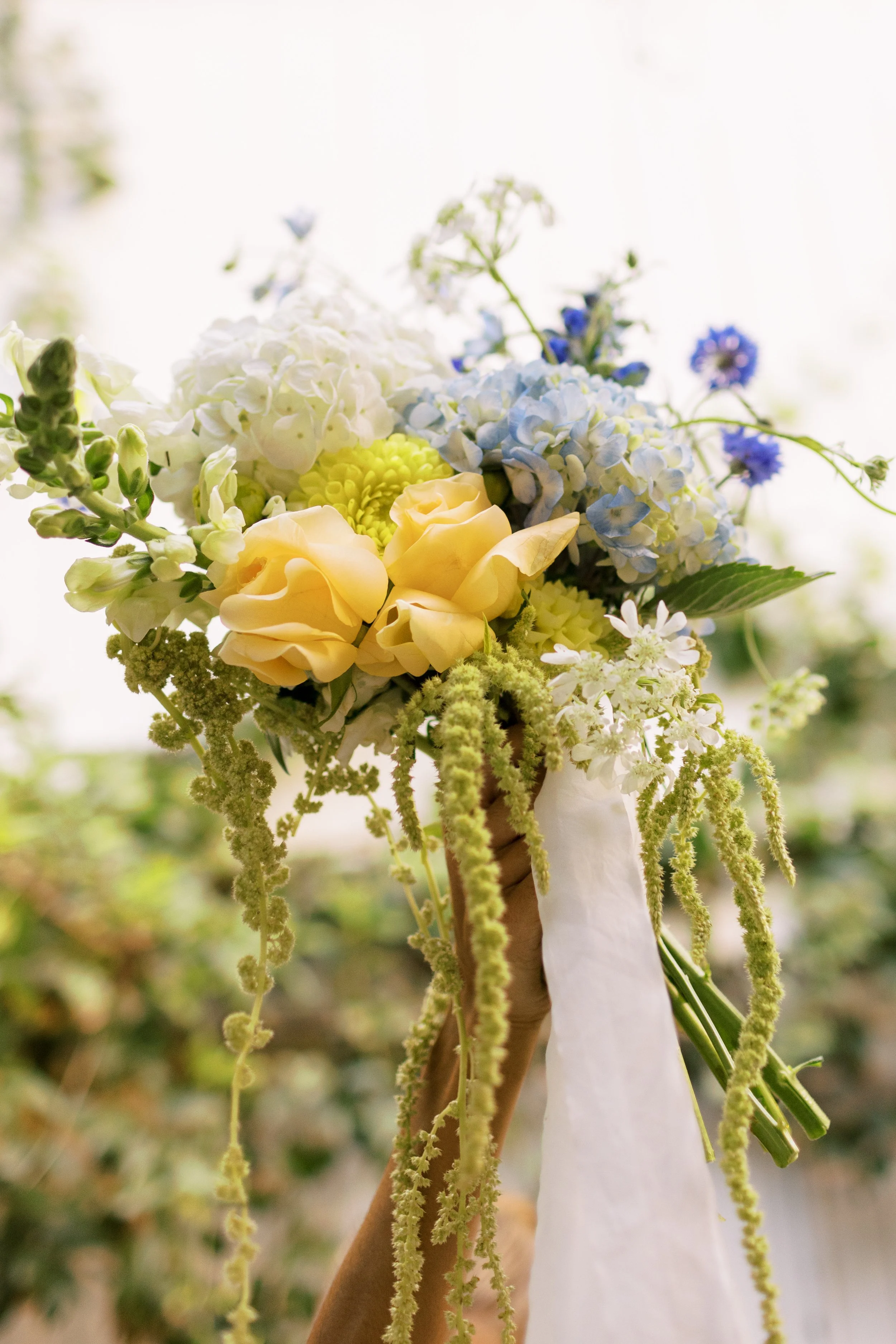 Hand holding a bouquet of flowers including yellow roses, white and blue hydrangeas, and white daisies, with a white ribbon tied around the stems.