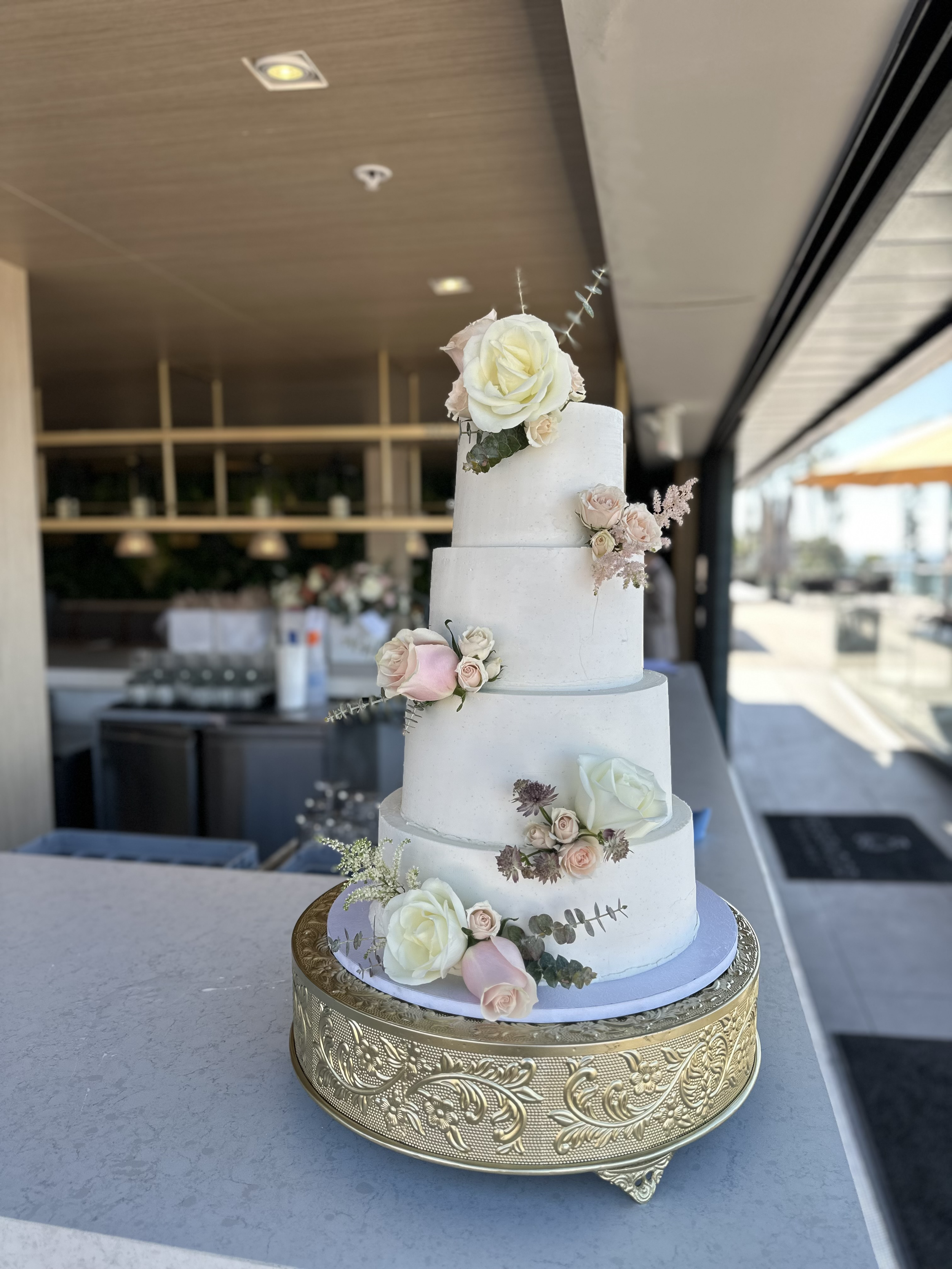 A four-tier white wedding cake decorated with pink and white roses and greenery, placed on a gold ornate cake stand, inside a venue with large windows and a view outside.