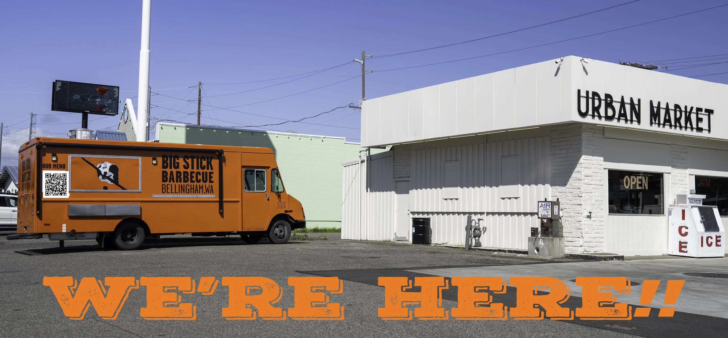 An orange food truck marked 'Big Stick Barbecue' parked outside a white building labeled 'Urban Market' on a sunny day with a clear blue sky.