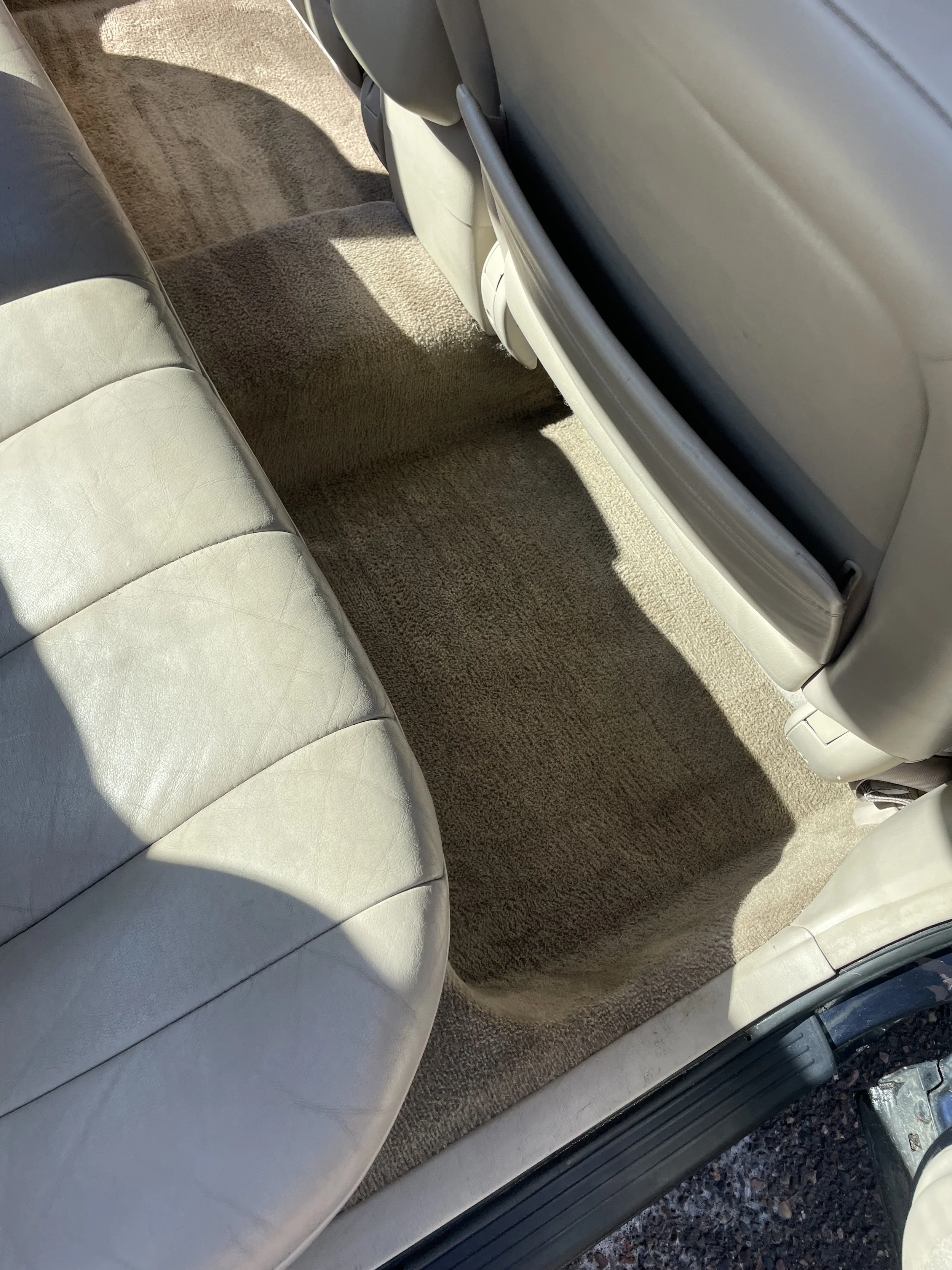 The back seat of a car with light-colored leather upholstery and a beige carpeted floor.