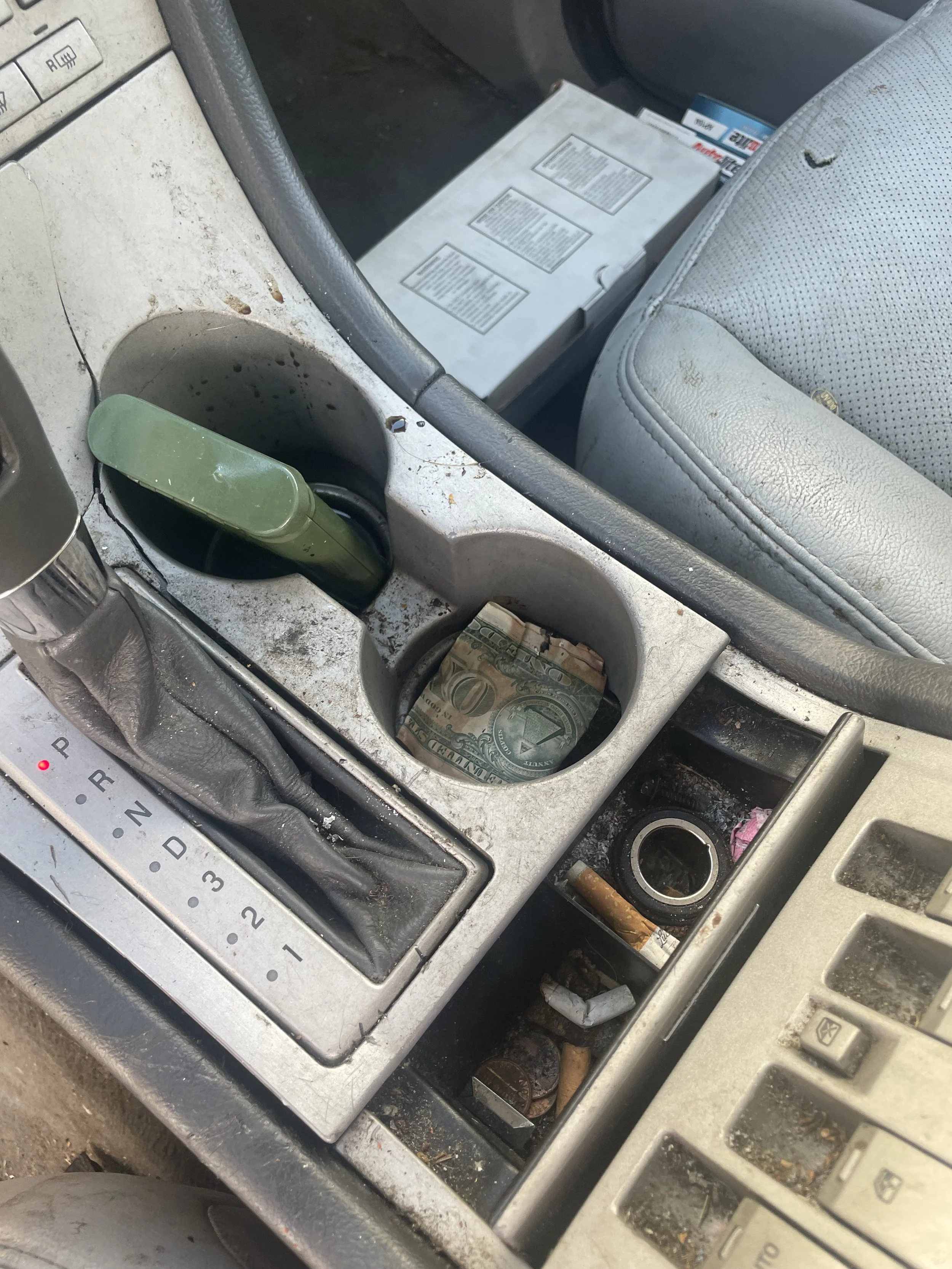 Inside a dirty car center console with a gear shift, containing a single dollar bill in a cup holder, a small cleaning stick, and some scattered cigarette butts and trash.