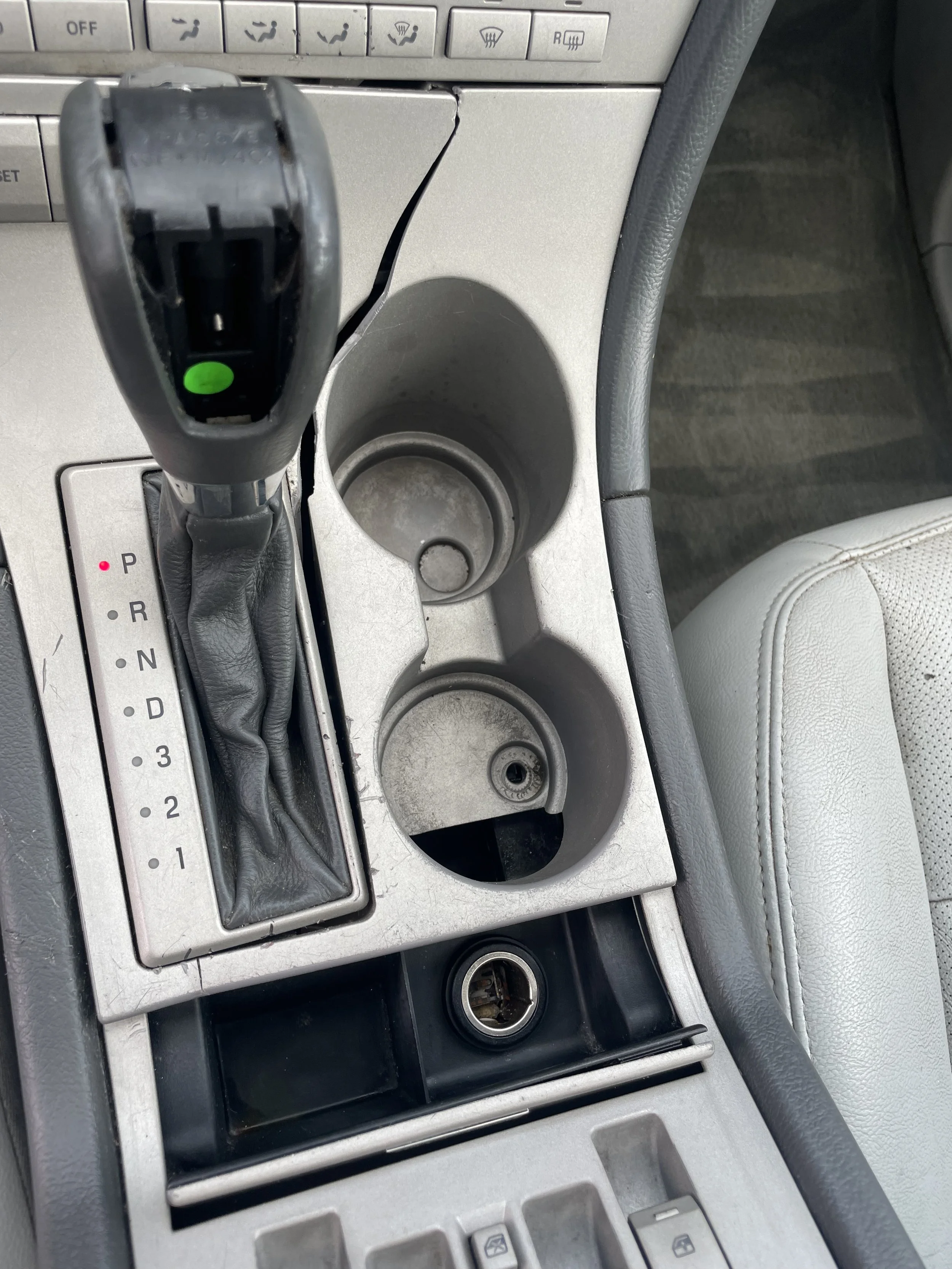Close-up of a car's center console with a gear shifter, cup holders, and missing cover for a cigarette lighter or power outlet.