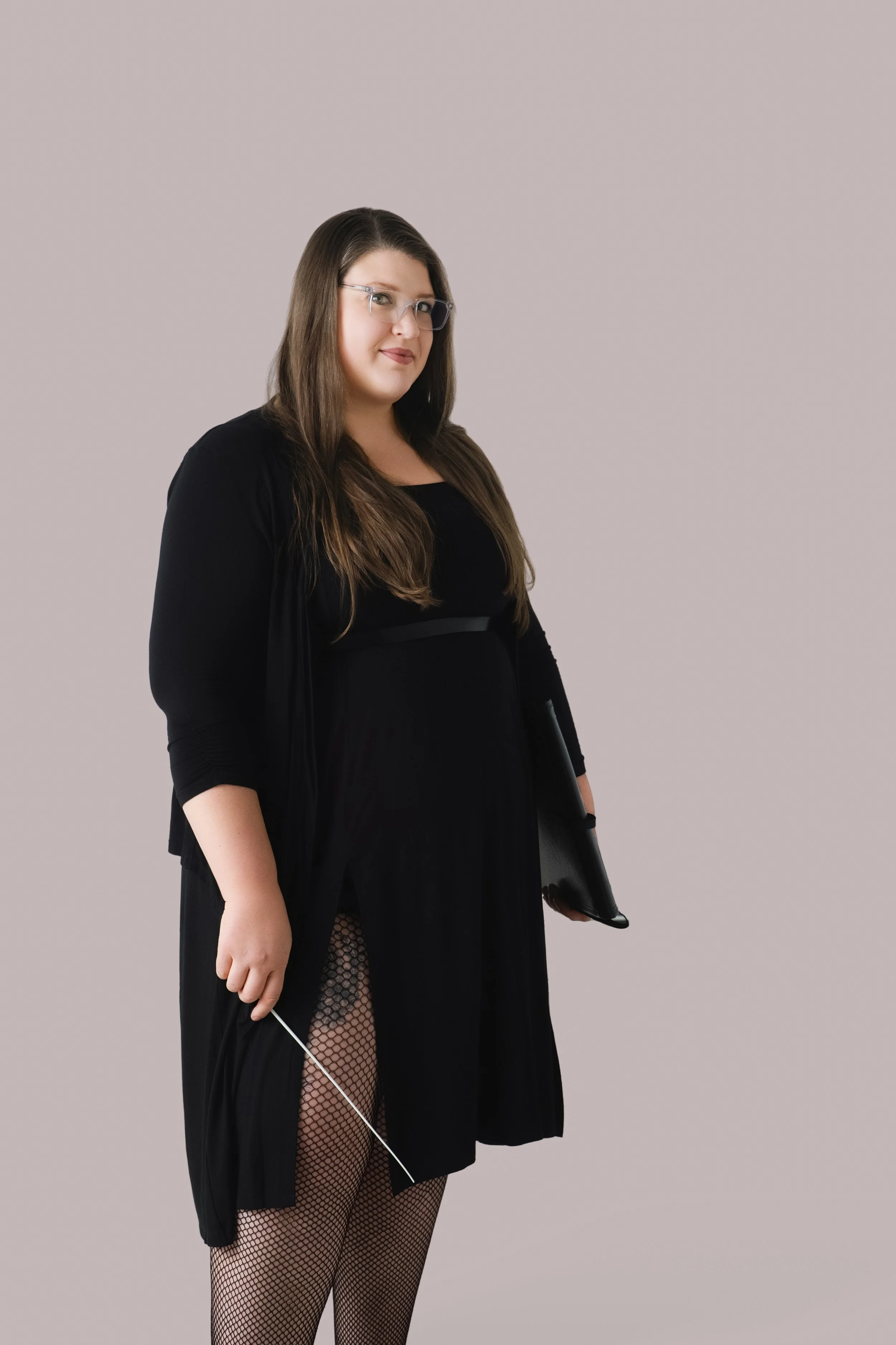A woman with long brown hair, glasses, wearing a black dress with fishnet stockings and a black cardigan, holding a black clutch bag, standing against a plain gray background.