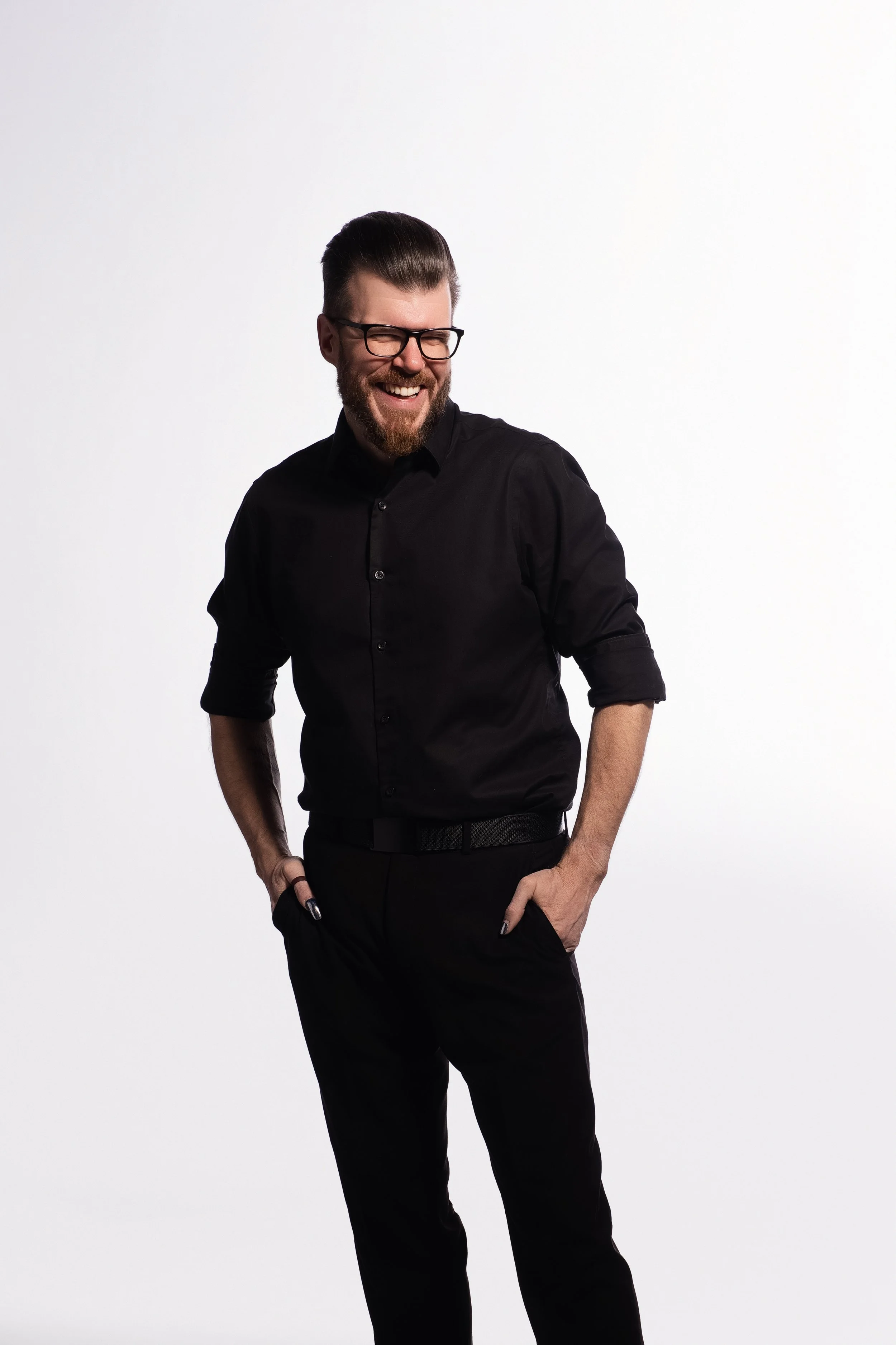 A man with dark hair and beard, wearing glasses, in a black shirt, smiles and looks to his left with his hands in his pockets against a plain white background.