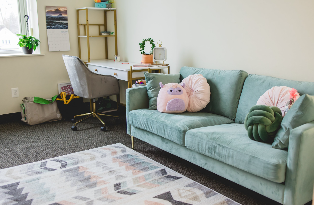 A living room corner with a mint green velvet sofa decorated with plush pillows, including a pink pig and a green knot pillow, next to a window. Behind the sofa, there is a small white desk with a gold frame, a gray chair, and various items on the desk. A calendar hangs on the wall, and a potted plant sits on the windowsill.