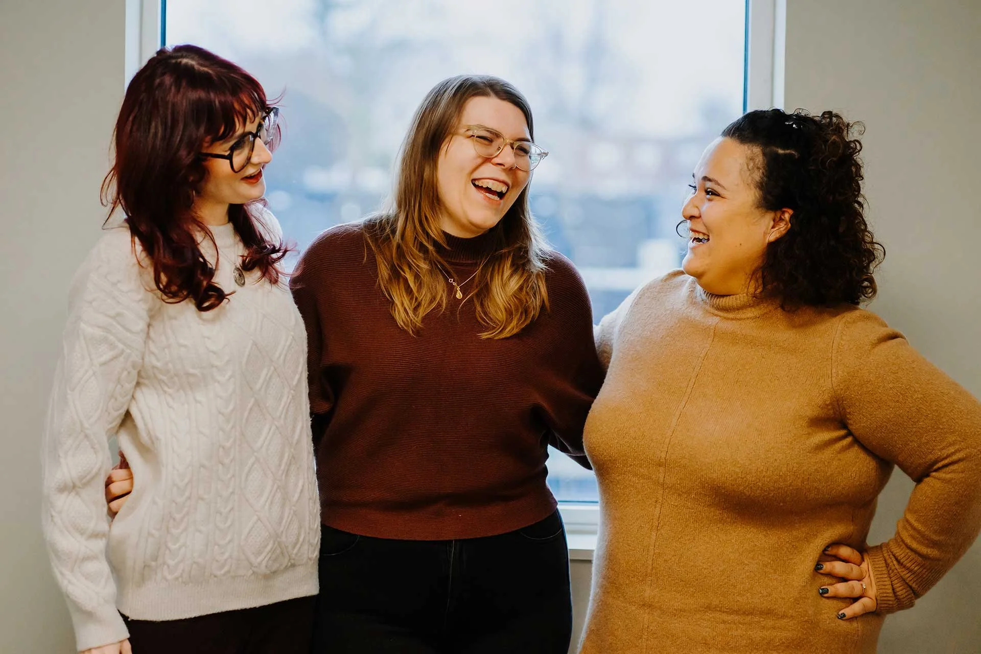 Therapists Faith Higgins, Kirstie Juenger and Eva Light standing together smiling and laughing in front of a window in their Bala Cynwyd office.