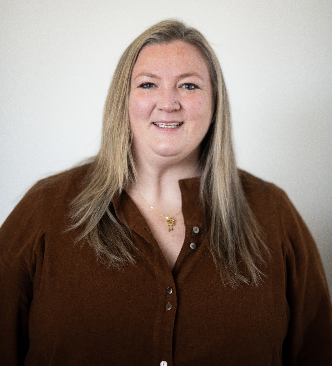 A woman with long blonde hair smiling at the camera, wearing a brown button-up shirt and a gold necklace.