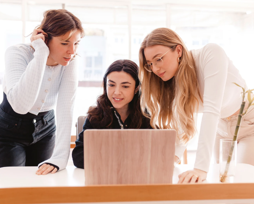Three women collaborating around a laptop in a bright office during hello DBT therapy.