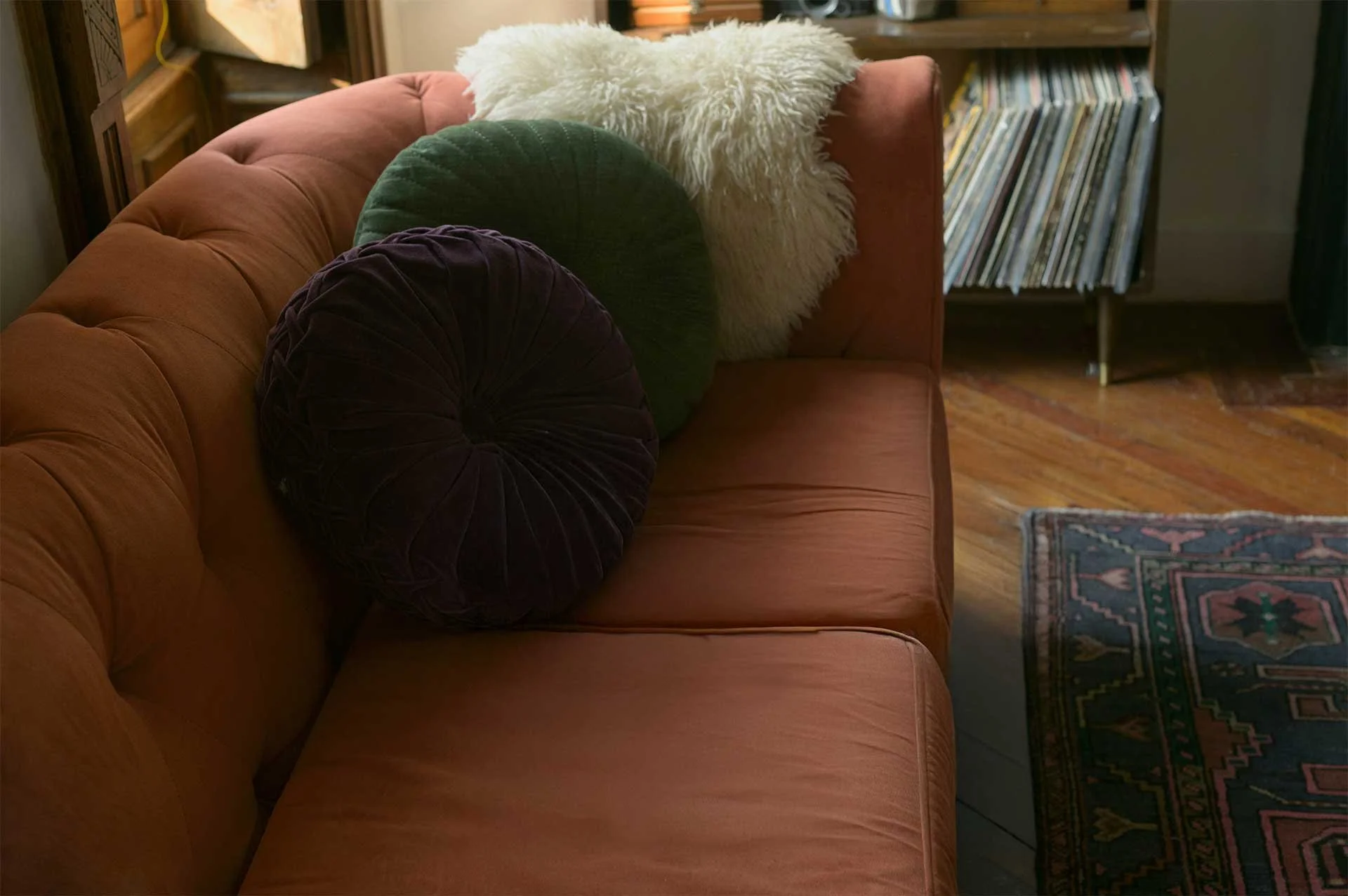 A rust-colored sofa decorated with assorted round pillows, including a white fluffy pillow, a green velvet pillow, and a dark purple velvet pillow, situated in a cozy living room with a wooden floor and a patterned rug.
