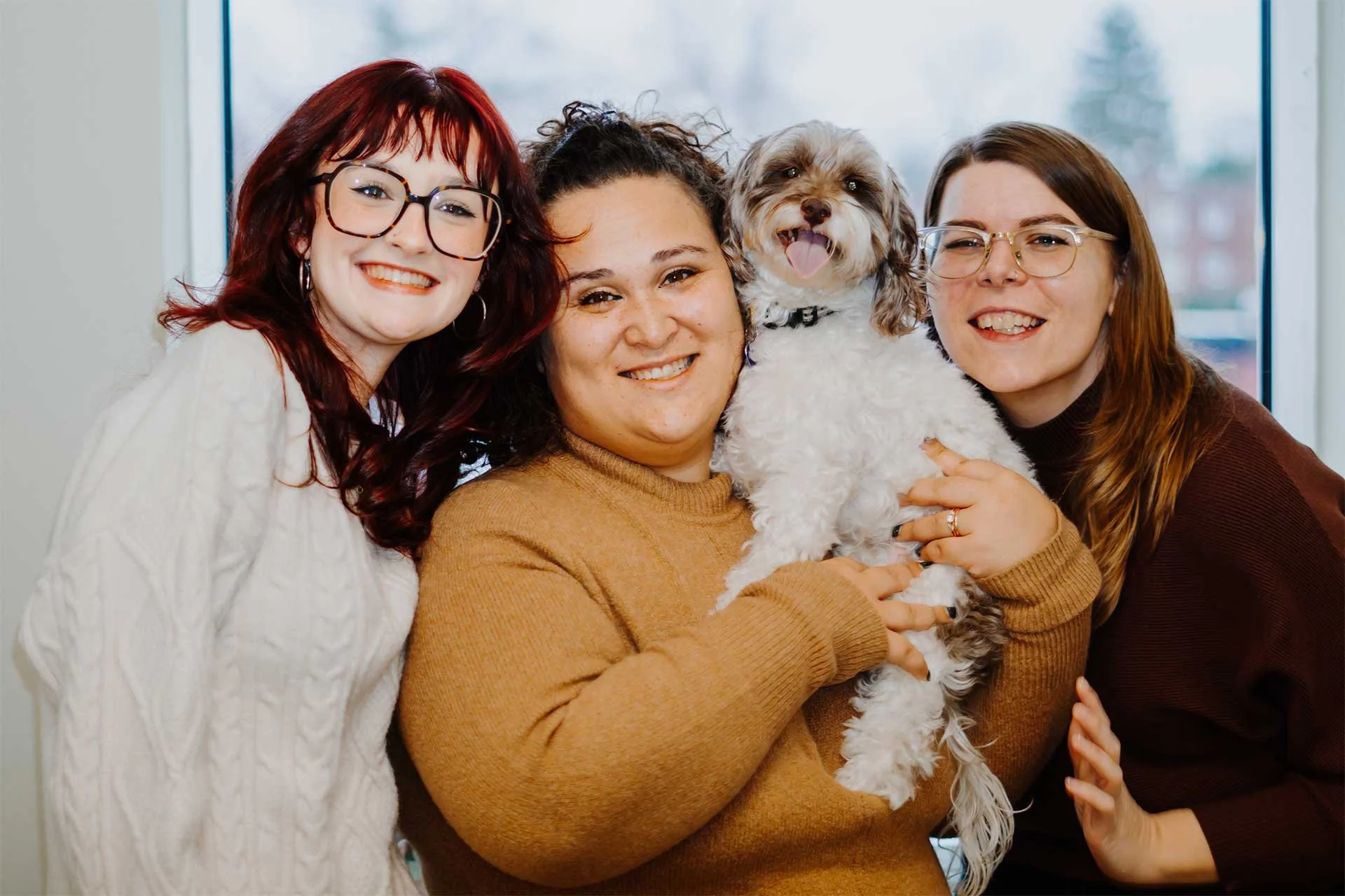 Therapists Faith Higgins, Kirstie Juenger and Eva Light smiling and posing together with therapy pup, Maisy in their brightly-lit office in Bala Cynwyd.
