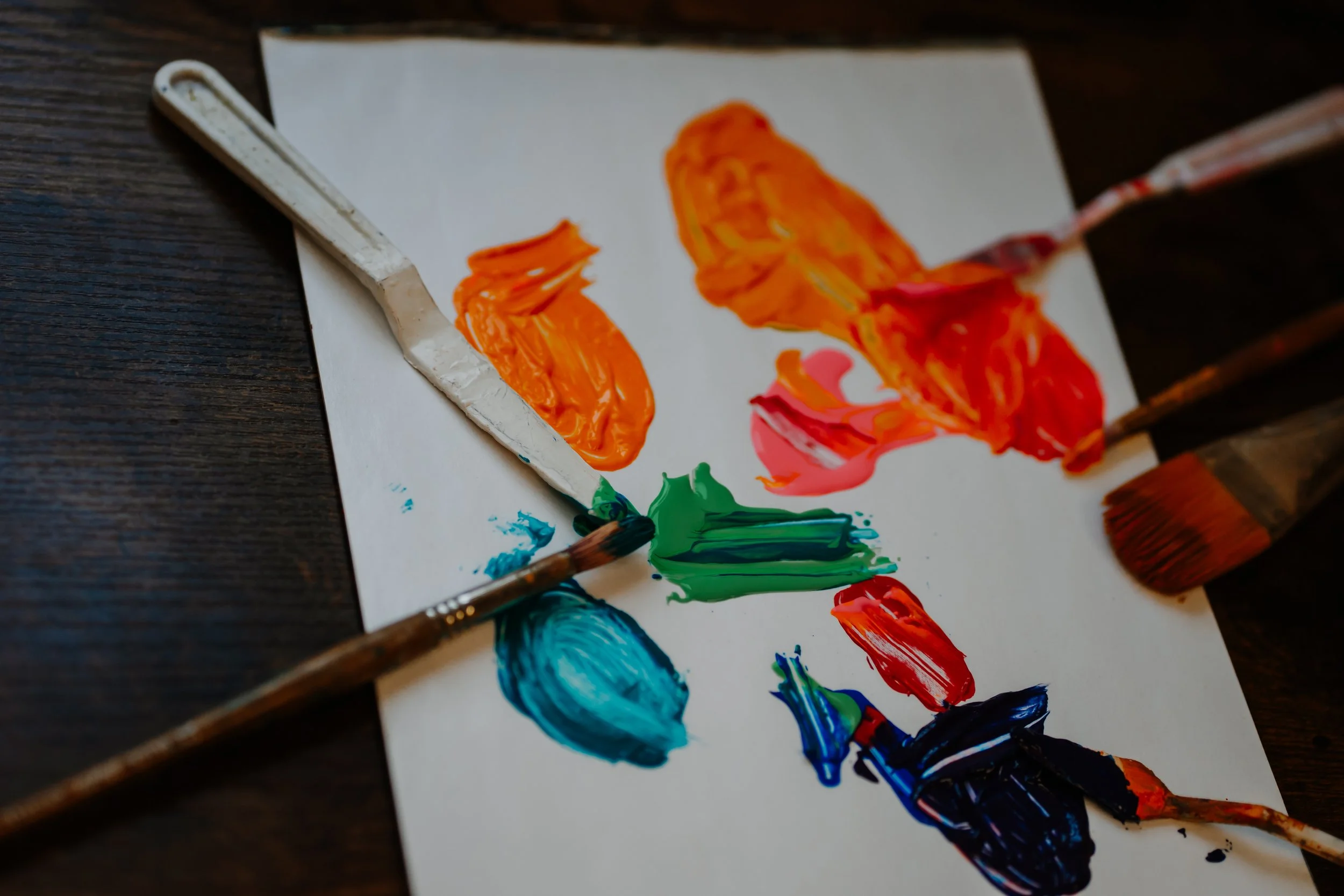 A painting palette with various dollops of brightly colored paint, including orange, pink, green, blue, red, and black, surrounded by paintbrushes on a white sheet of paper and a dark wooden surface.