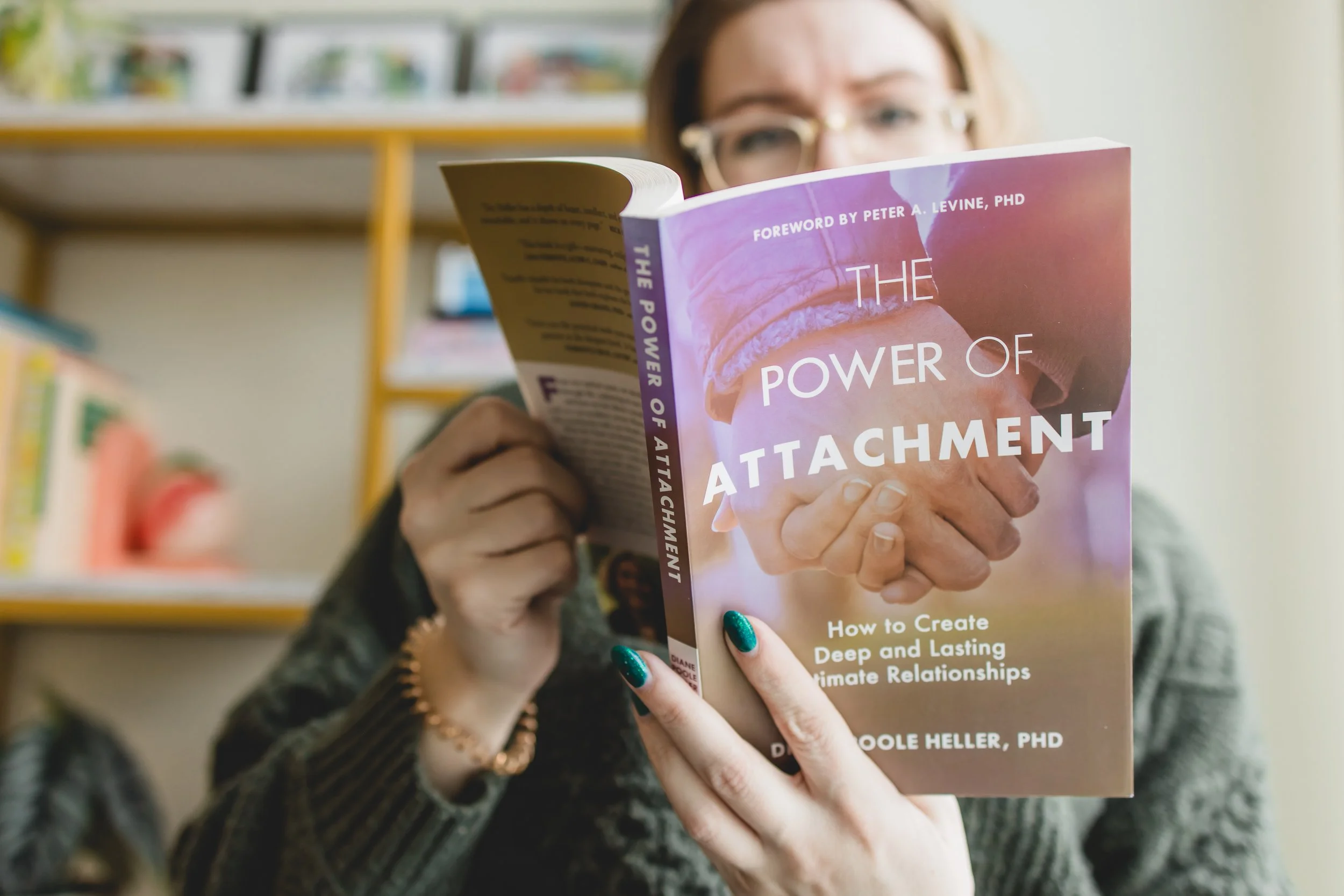 A woman with glasses and a sweater is reading a book titled 'The Power of Attachment' in a room with shelves of books in the background.