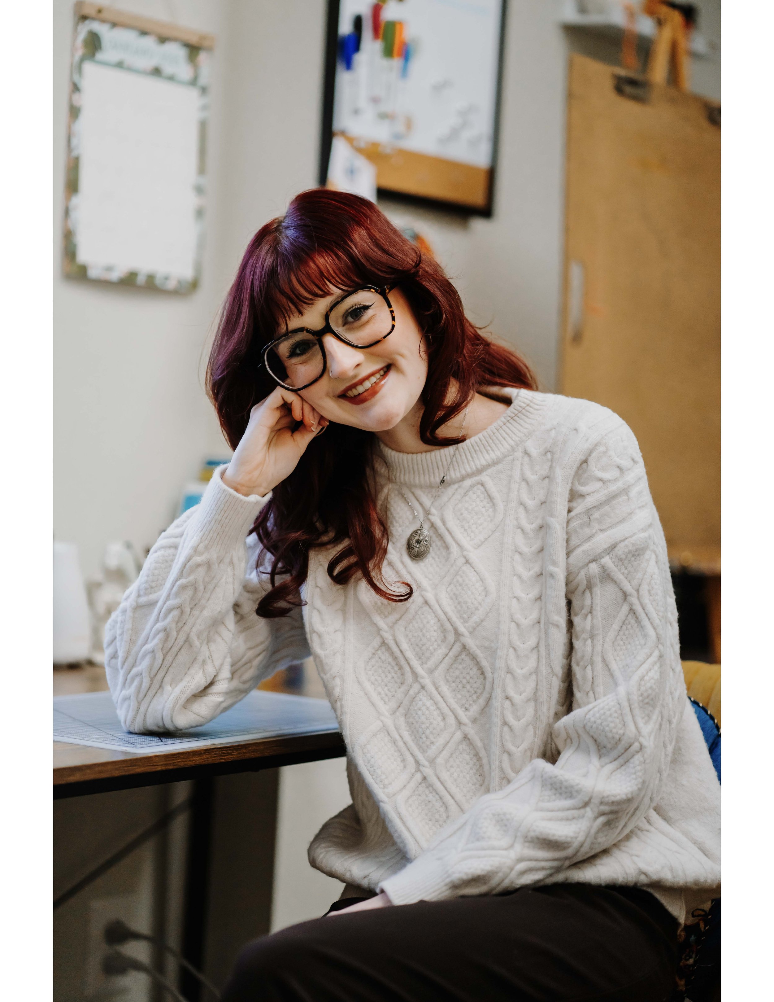 A young woman with red hair, glasses, and a white cable-knit sweater is sitting at a desk, smiling with her head resting on her hand in an indoor setting. There are bulletin boards and a wooden cabinet in the background.