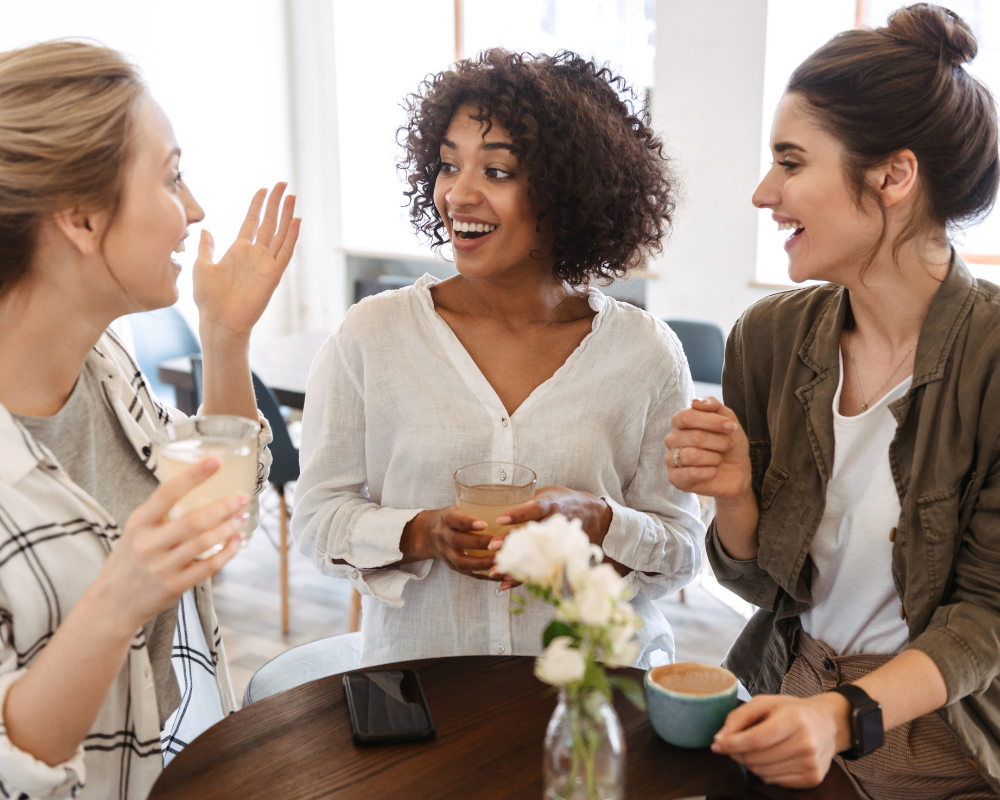 Three women smiling and talking while standing around a kitchen table with coffee mugs and a vase of flowers.