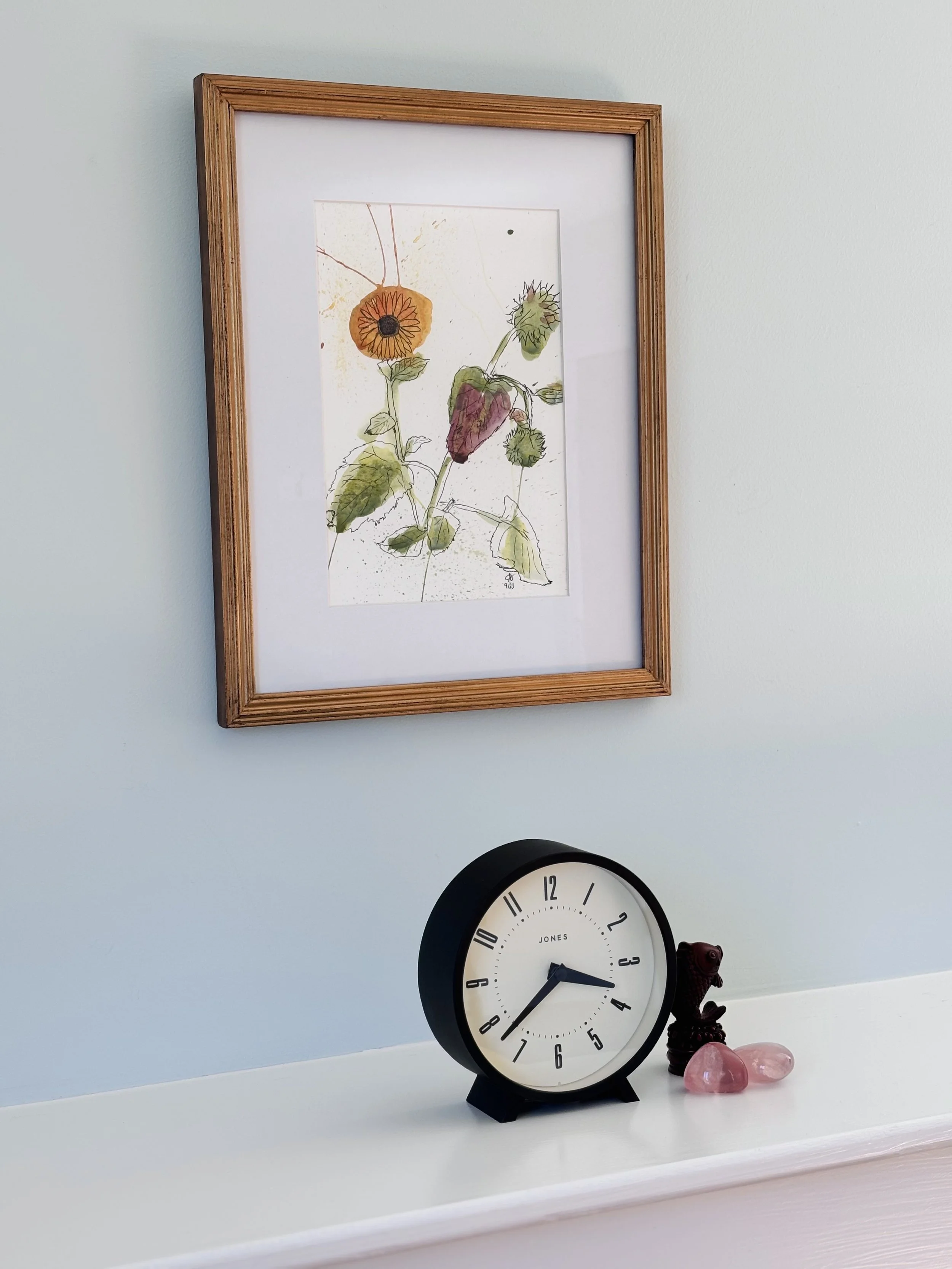 Wall clock with black frame and white face, placed on a white surface, with small decorative stones and figurines beside it. Framed artwork of watercolor flowers hangs on the wall above.