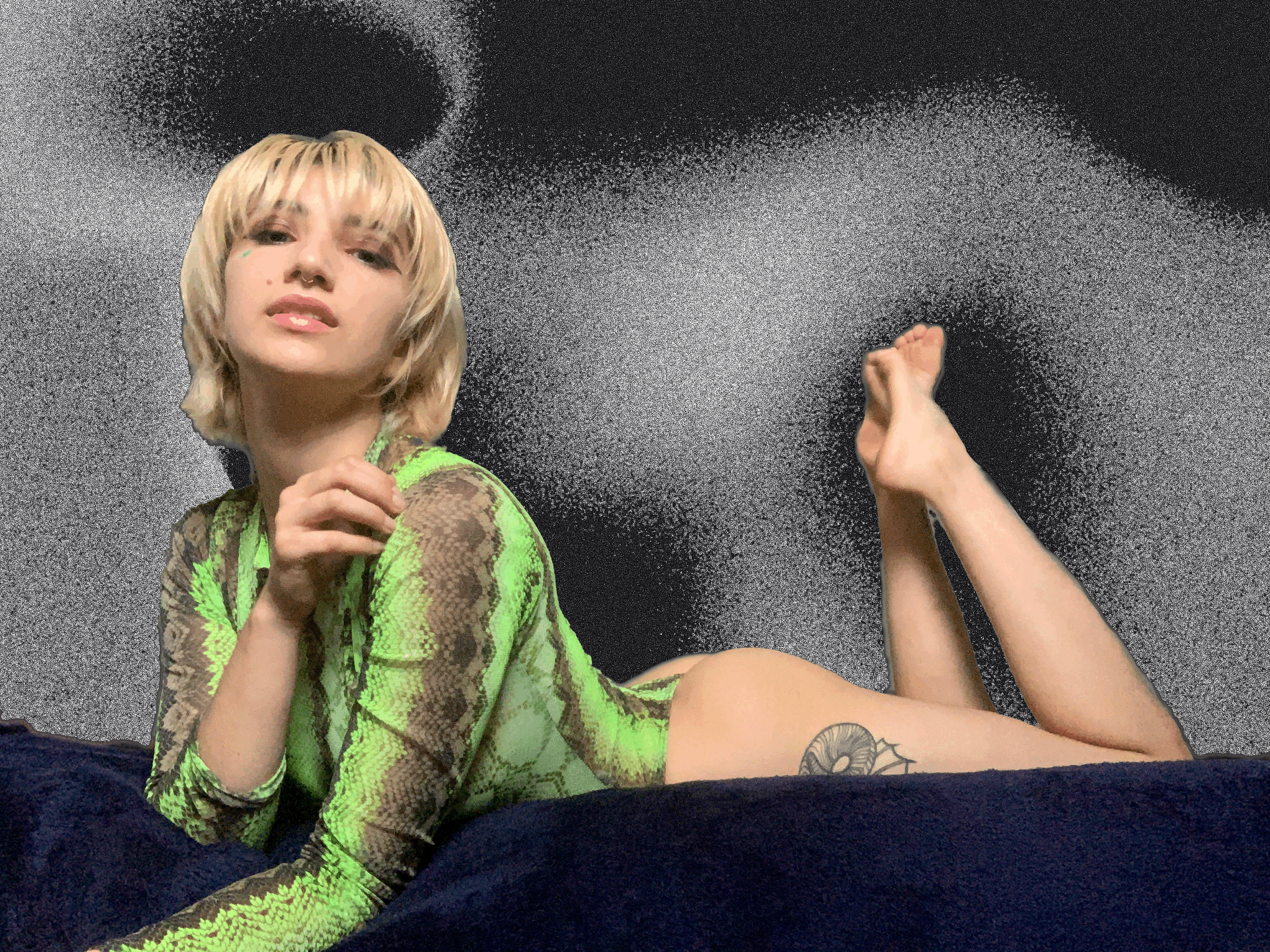 A woman with blonde hair laying on her stomach on a dark bed, wearing a neon green and brown snake print top, with a tattoo of a snail shell on her thigh, against a dark textured wall background.