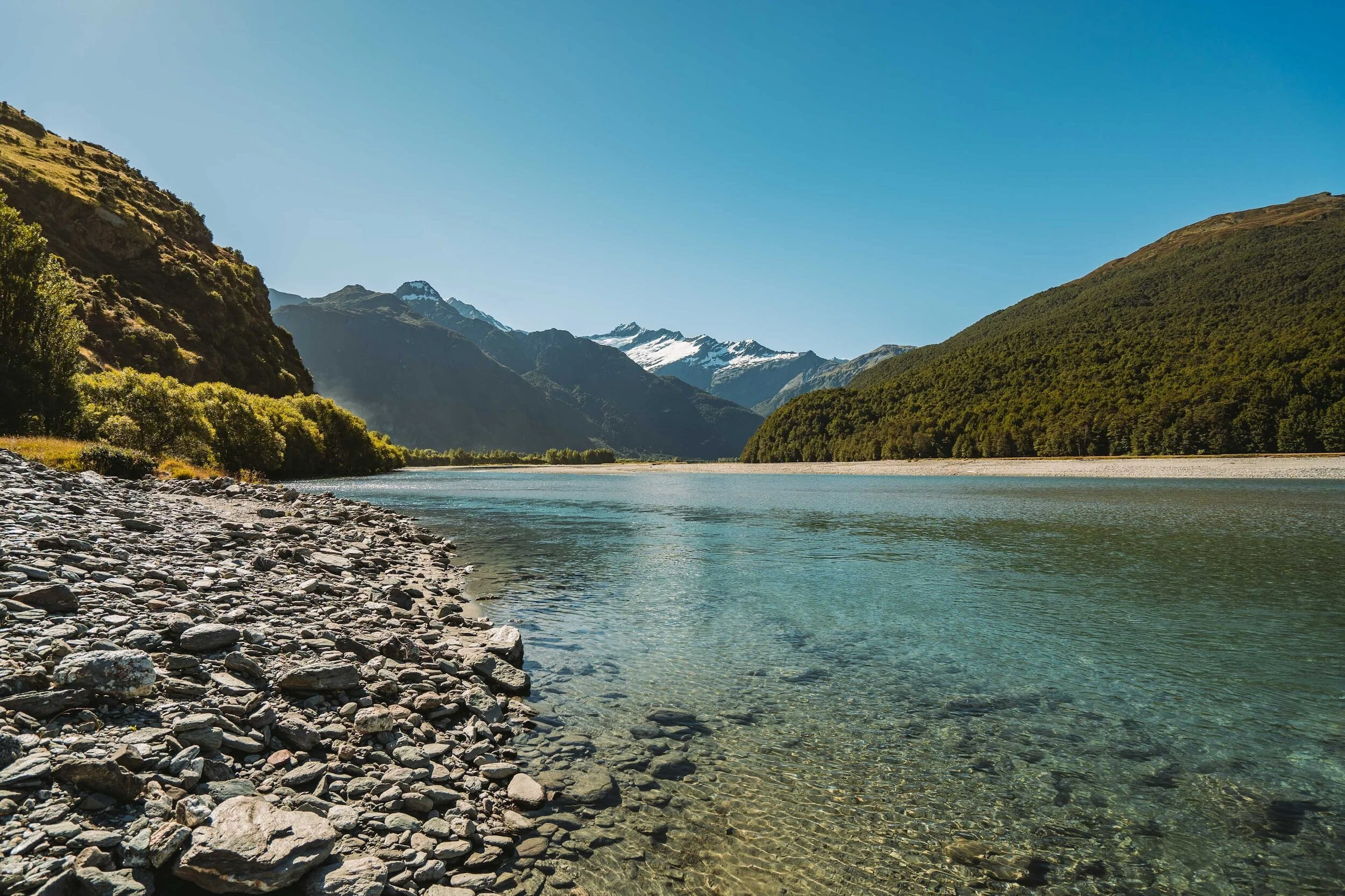 A clear river flowing through a mountainous landscape with snow-capped peaks in the background and rocky shoreline in the foreground.