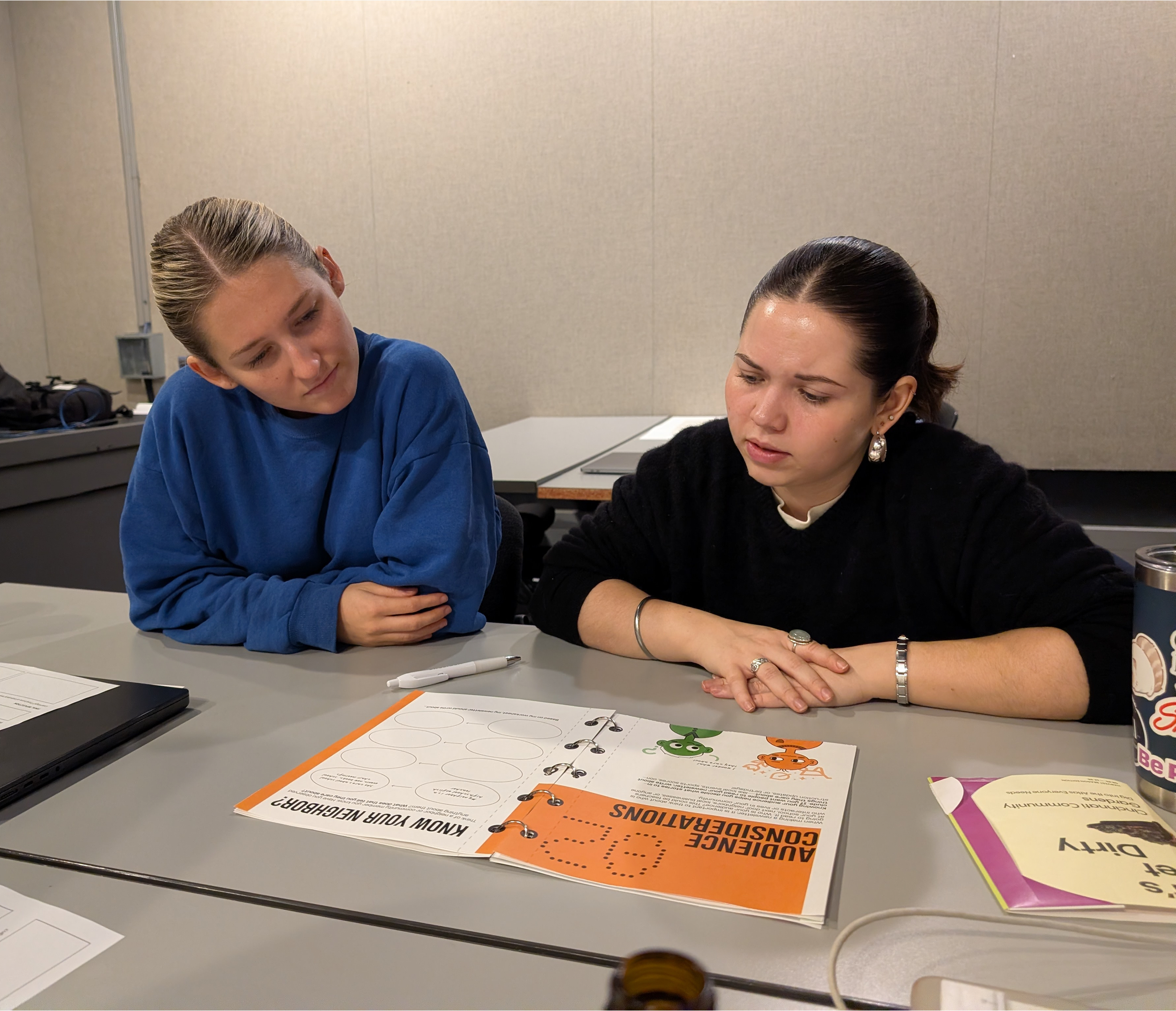 Two women sit at a desk looking at papers and a workbook with colorful cartoon characters, in a room with beige walls. One woman wears a black sweater and silver jewelry, the other wears a blue sweatshirt.