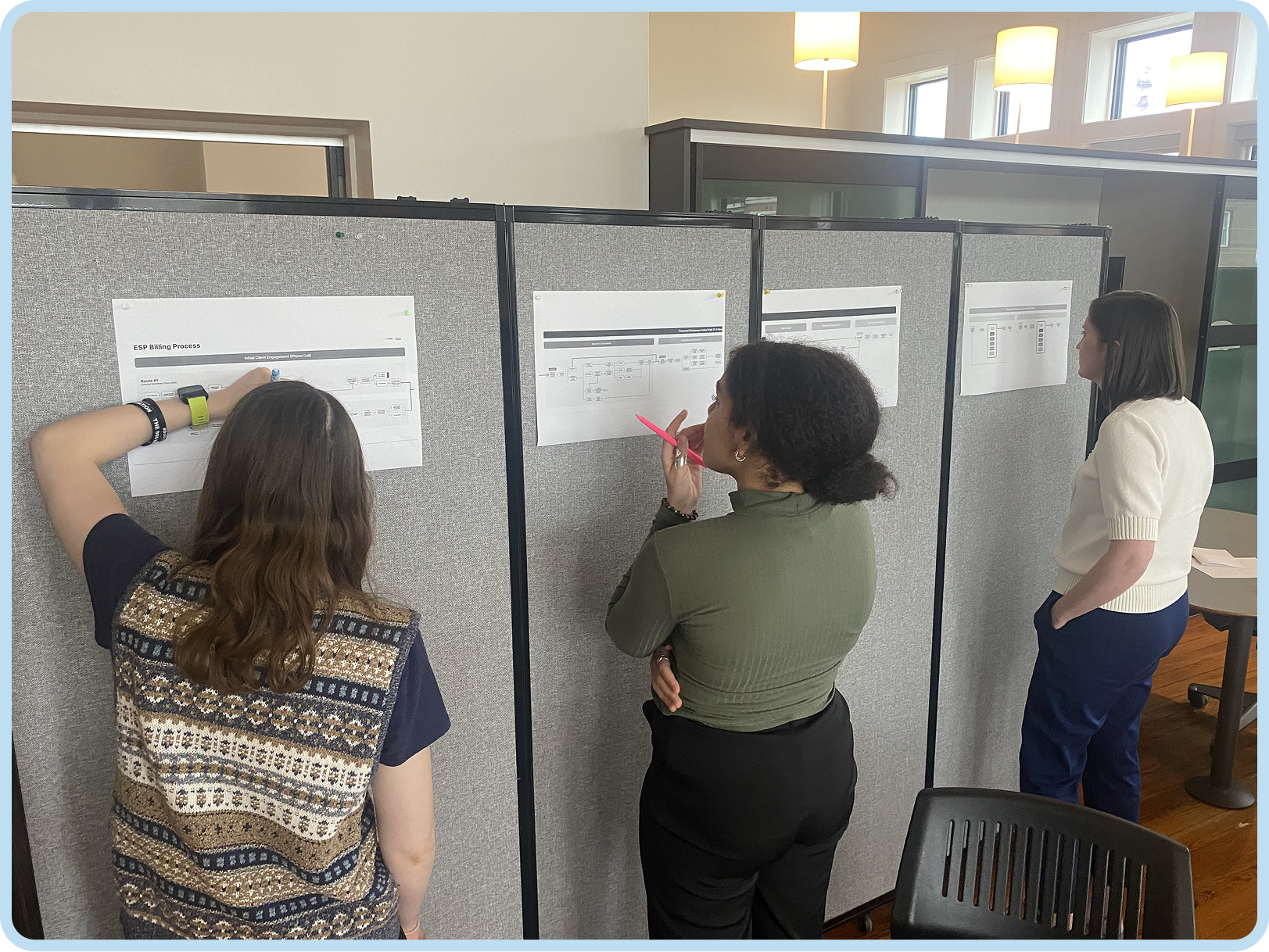 Three women standing and looking at posters with diagrams on gray partition panels in an office or conference room.