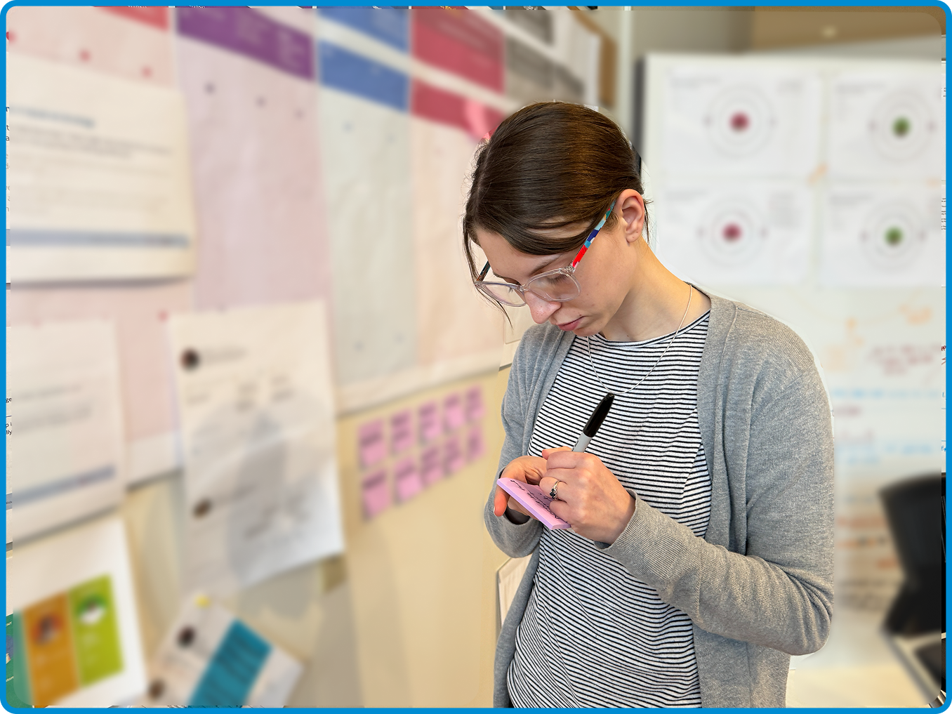 Woman with glasses writing on a pink notepad in an office or classroom with charts and papers on the wall.