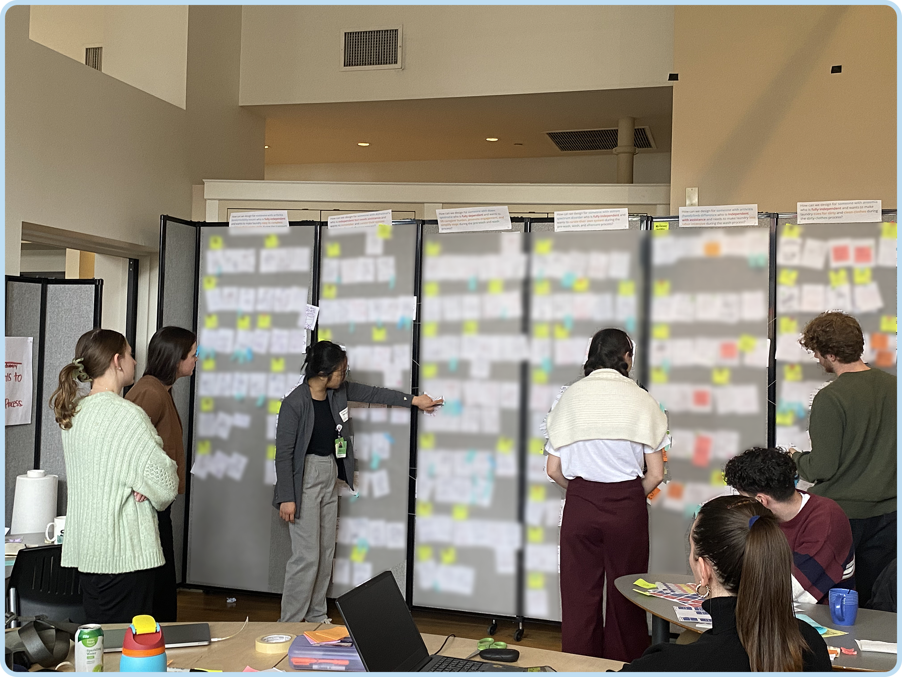 Several people standing and sitting in front of a large board with papers and notes displayed. The setting appears to be a conference or workshop, with tables, a laptop, and various office supplies visible.