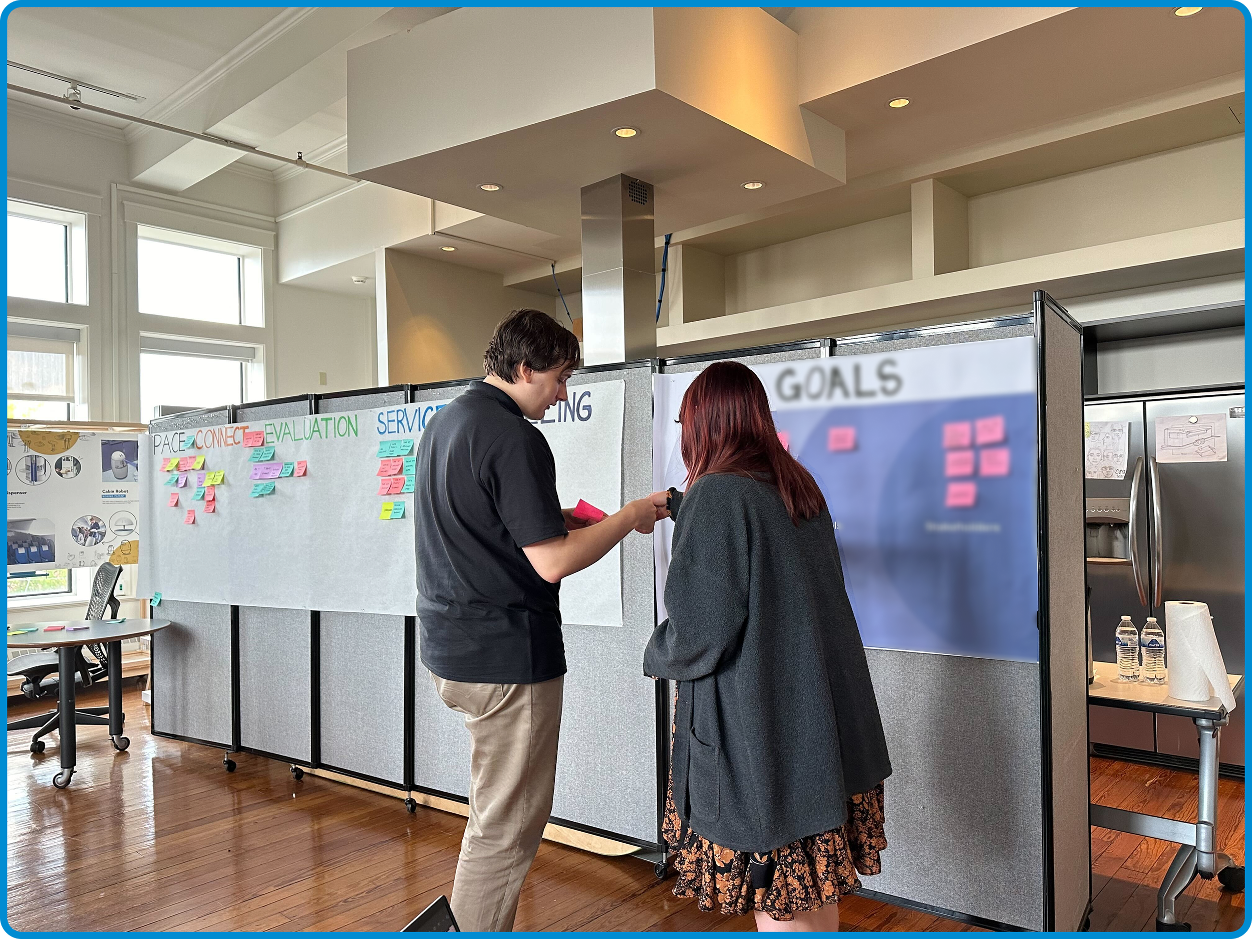 Two people standing and talking in front of a display board with sticky notes, with windows in the background and a wooden floor.