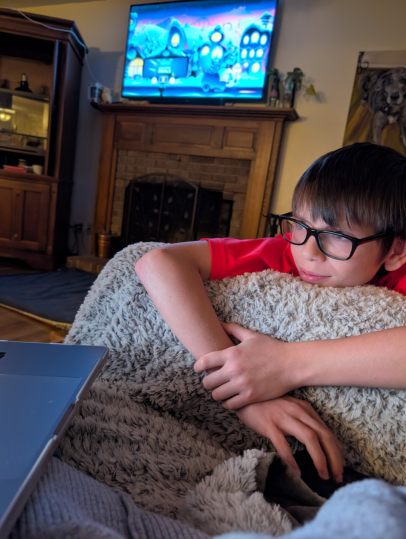 A young boy with glasses, wearing a red shirt, is lying on a plush blanket, resting his head on his arm, and watching a television.
