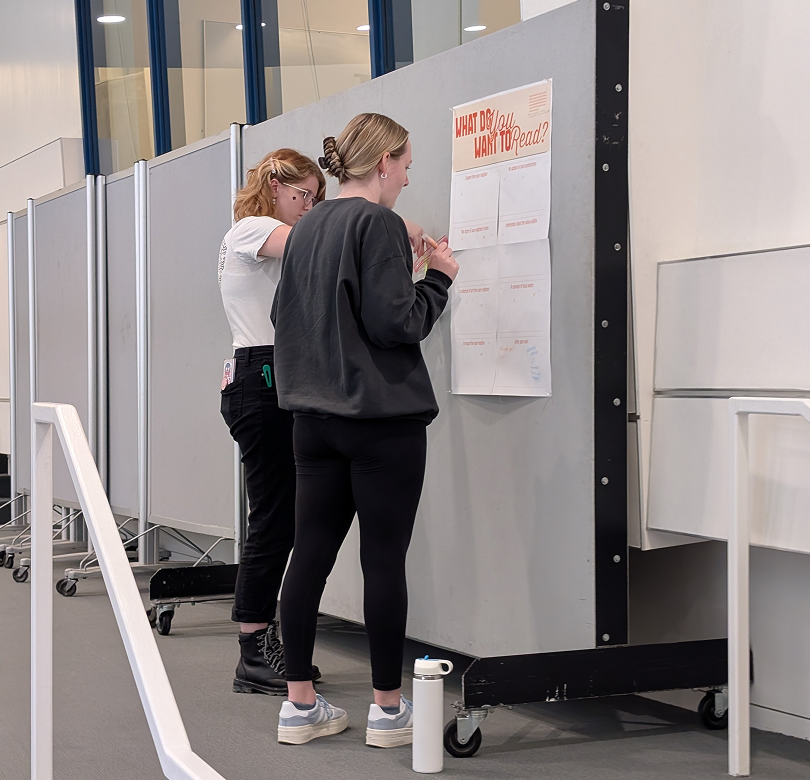 Two women are standing at a bulletin board, writing on a paper with the heading 'What Do You Want To Read?' in a public indoor space.