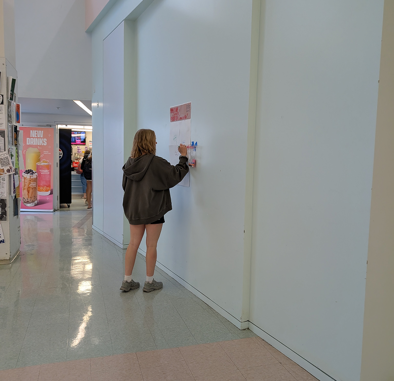 A person with red hair, wearing a dark hoodie, shorts, and hiking shoes, stands facing a white wall in a shopping mall, writing or drawing on a poster board mounted on the wall.