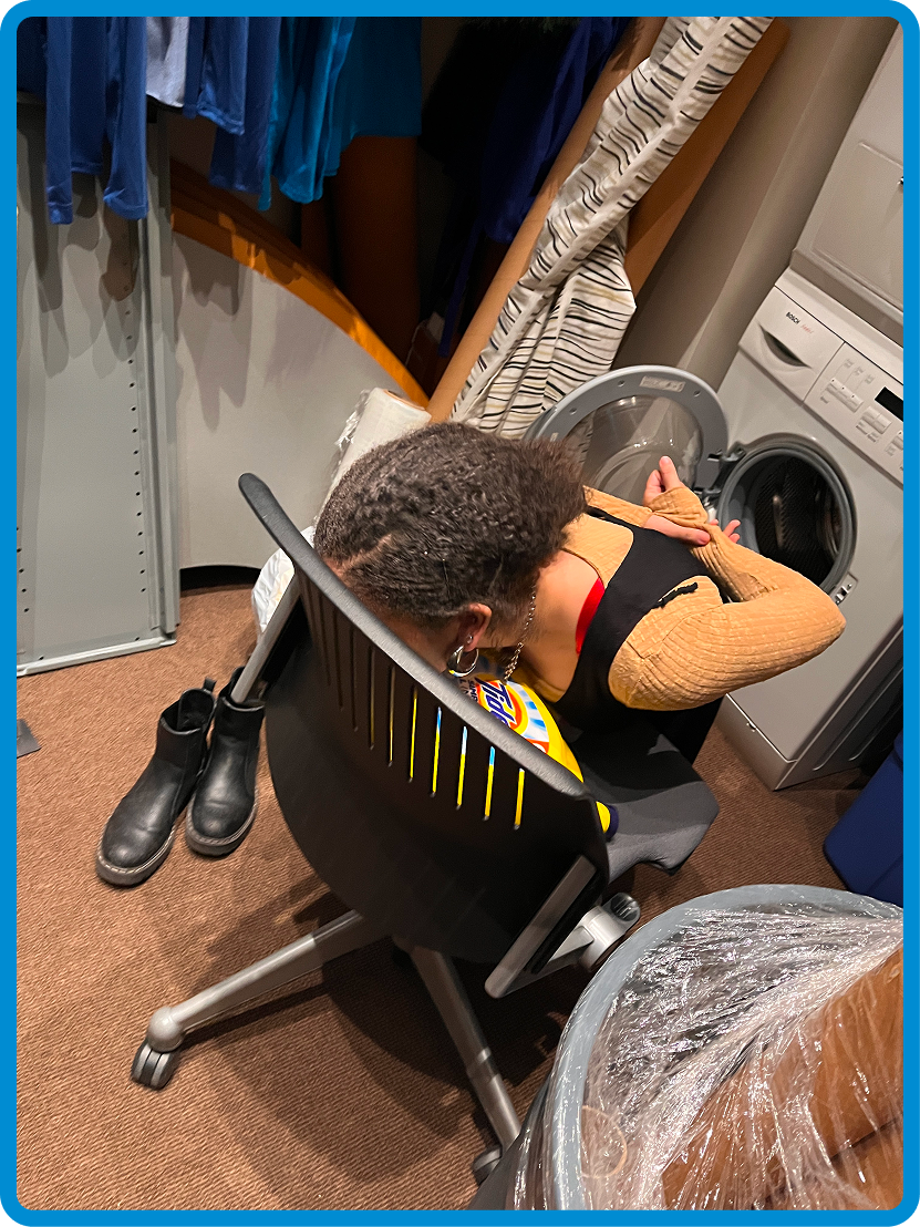 A person with short hair and earrings, wearing a black sleeveless top and orange long-sleeve shirt, is leaning into a washing machine in a laundry room.