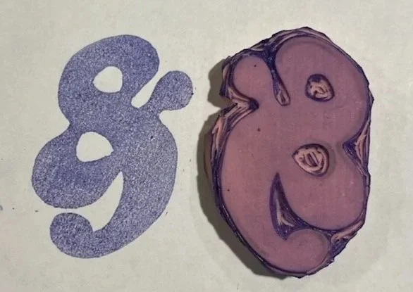 A linoleum block print with a purple or brownish hue, next to a printed shape resembling a stylized ampersand, with a textured surface.