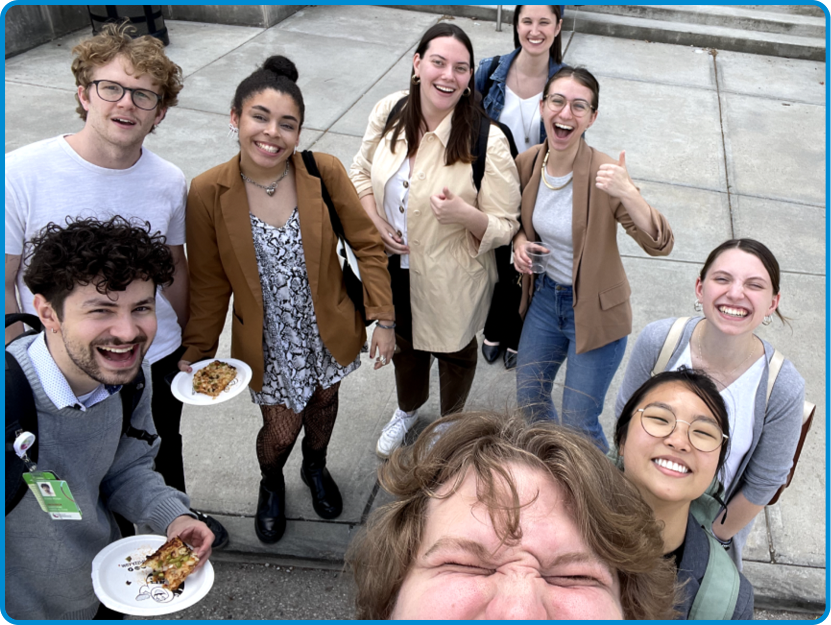 A group of diverse young adults smiling and posing for a selfie outdoors on concrete steps. Some are holding plates with food, and one person gives a thumbs-up gesture.
