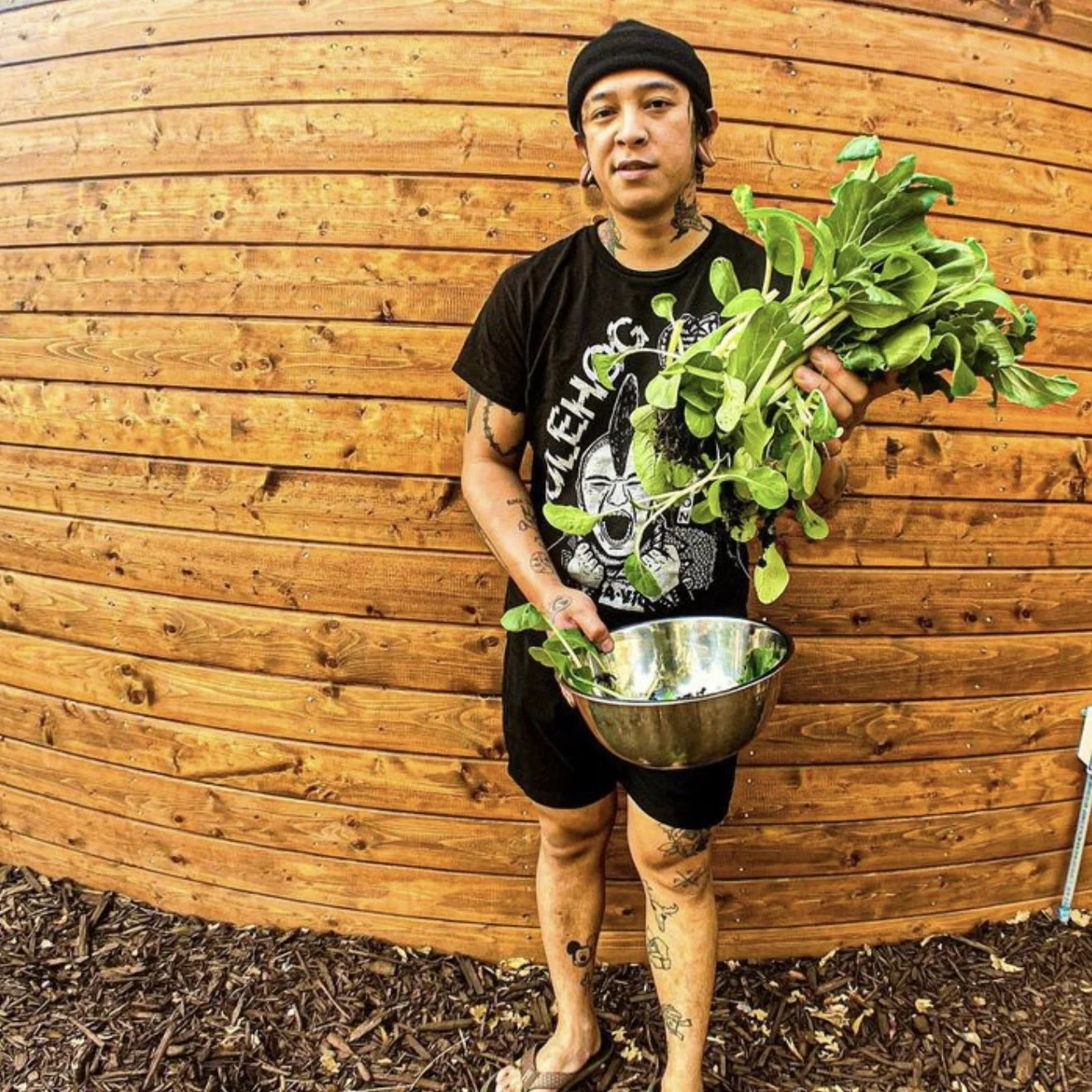 Ronnie Apolo pictured holding a very large bunch of freshly harvested greens and a stainless steel bowl. He is wearing black graphic tee, a beanie, and shorts, and is standing outside in front of plank wood wall.