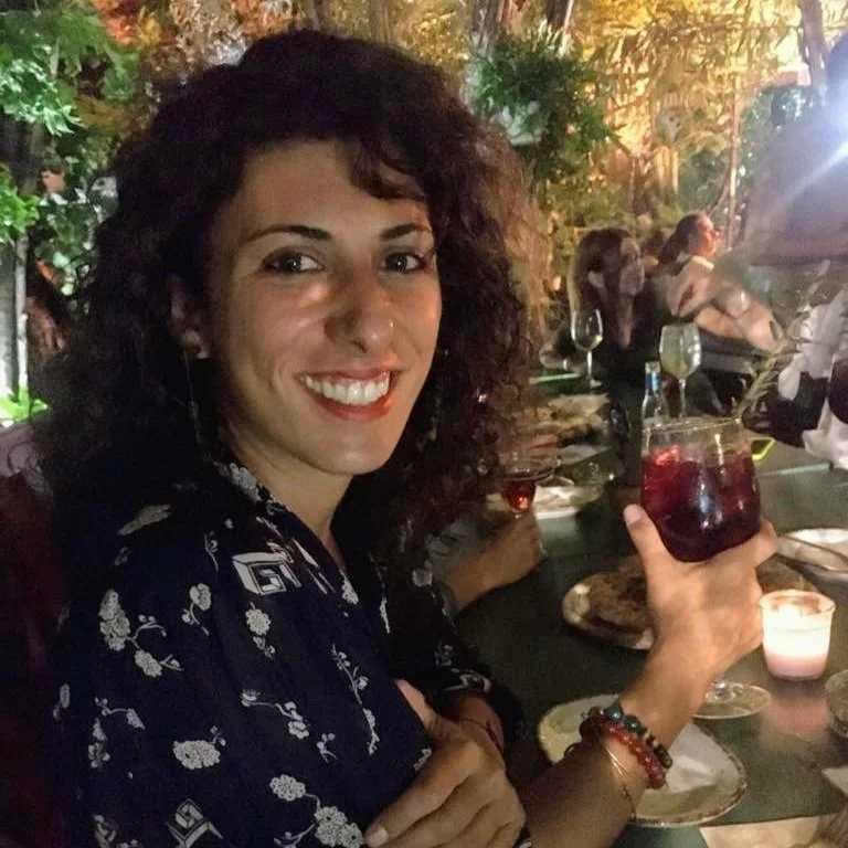 Simona Vallone is smiling broadly and looking to the camera while holding a red hued cocktail. She has her dark curly hair down and is wearing a black collared shirt with white graphic pattern. She is sitting at a counter in a restaurant.