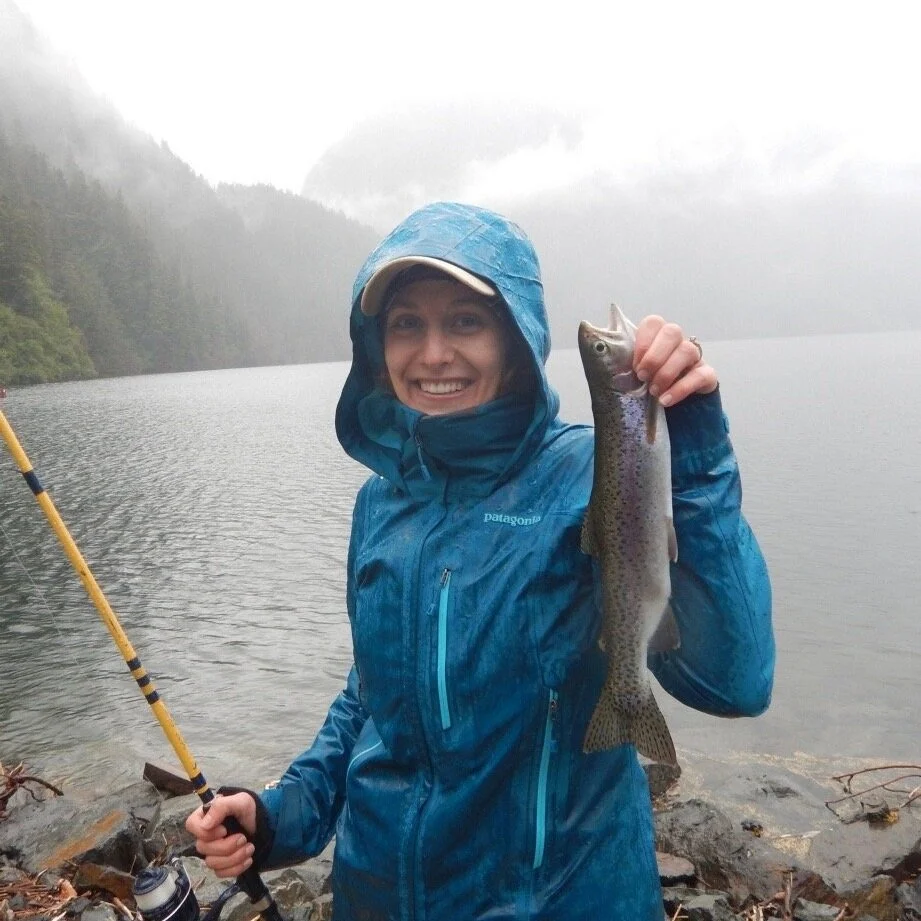 Salle Madison smiling widely at the camera wearing bright teal rain gear. It is rainy and she's holding her catch, a salmon, in one hand, a pole in the other hand, and standing on the edge of an Alaska waterway.