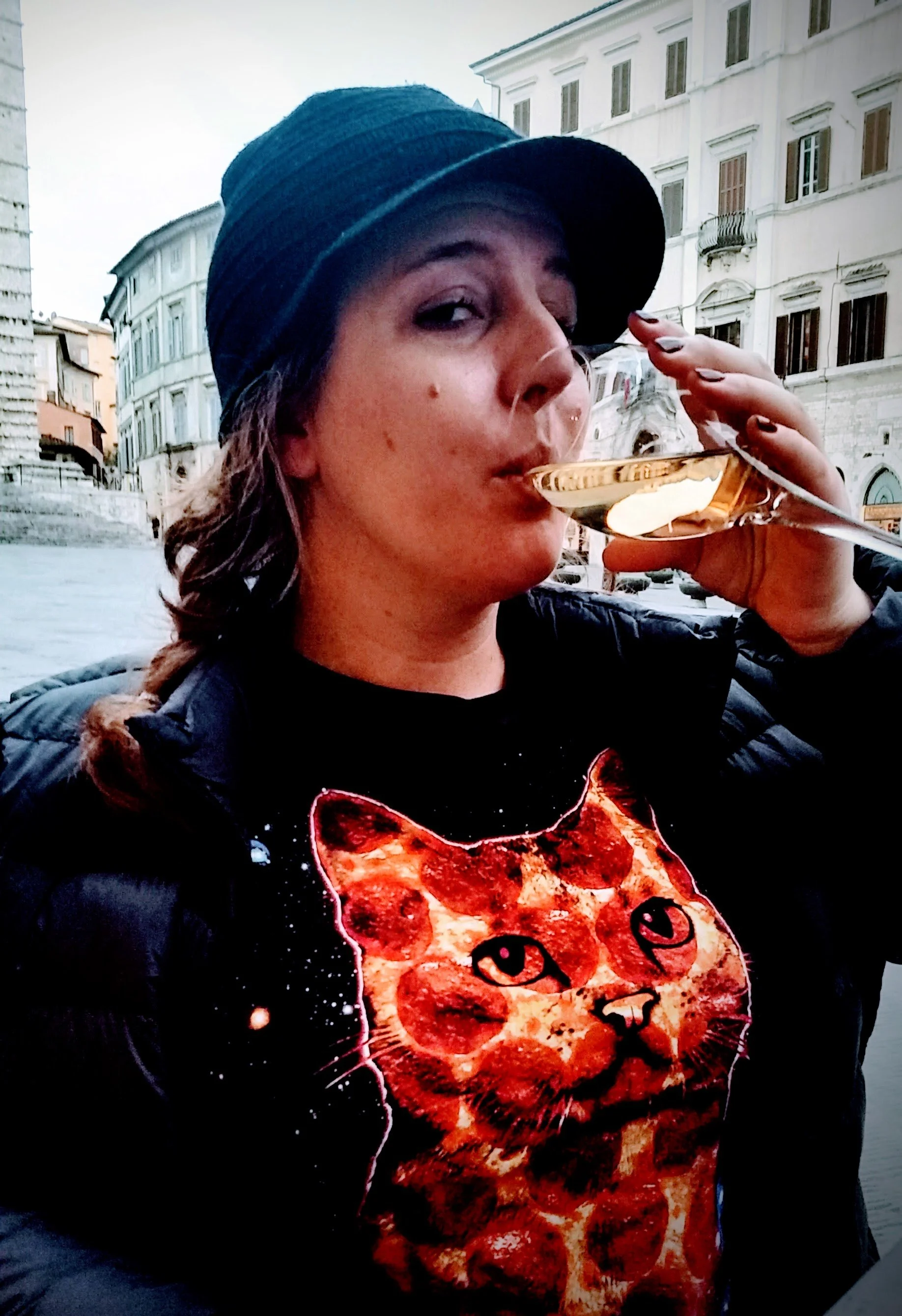 Kali Mulligan profile view while sipping a glass of rose wine and wearing a dark shirt with an image of a cat shaped pizza. A city neighborhood is in the background.