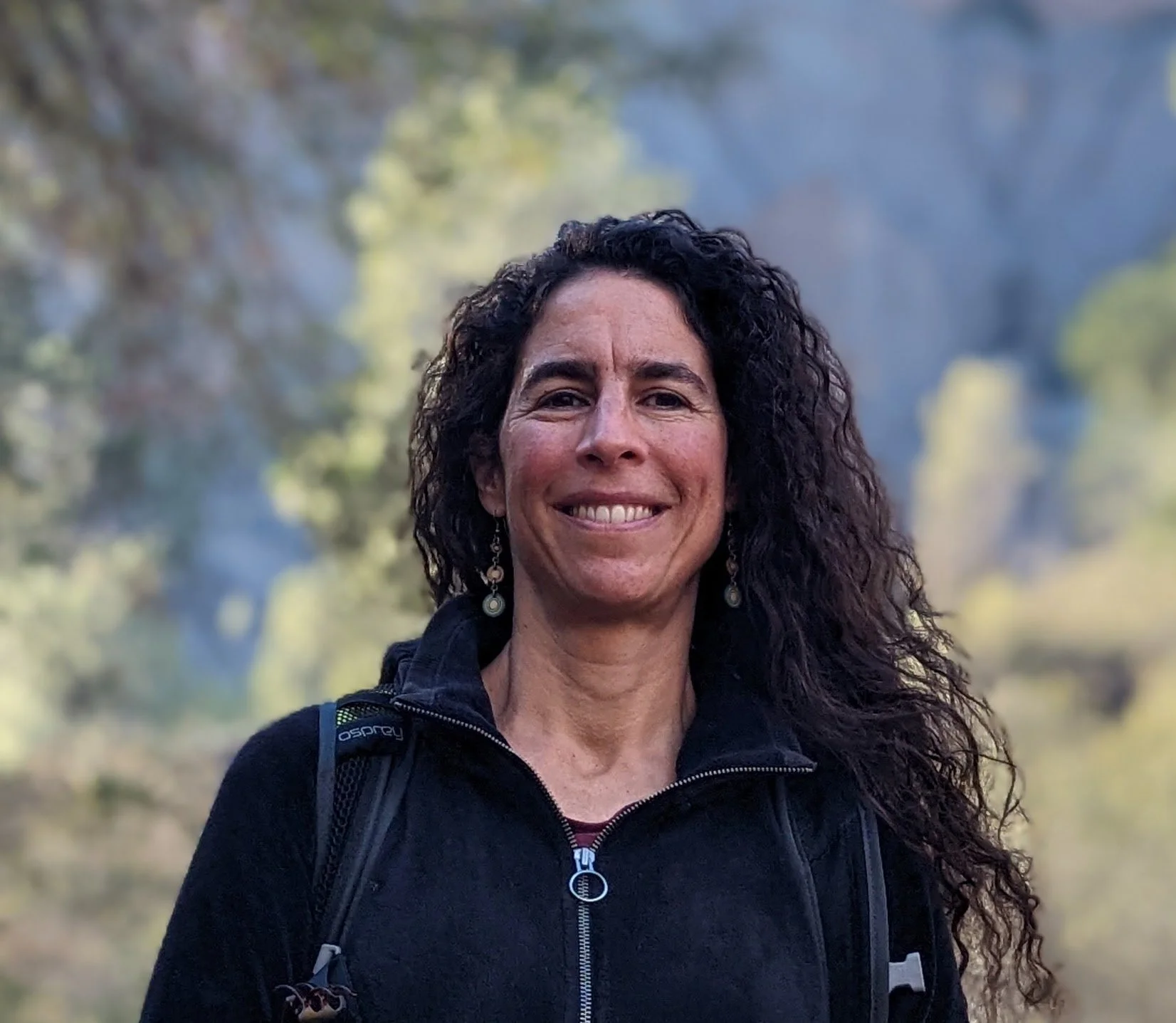 Adriana Telias shown with long dark curly hair wearing black hoodie with an outdoor scene in background.