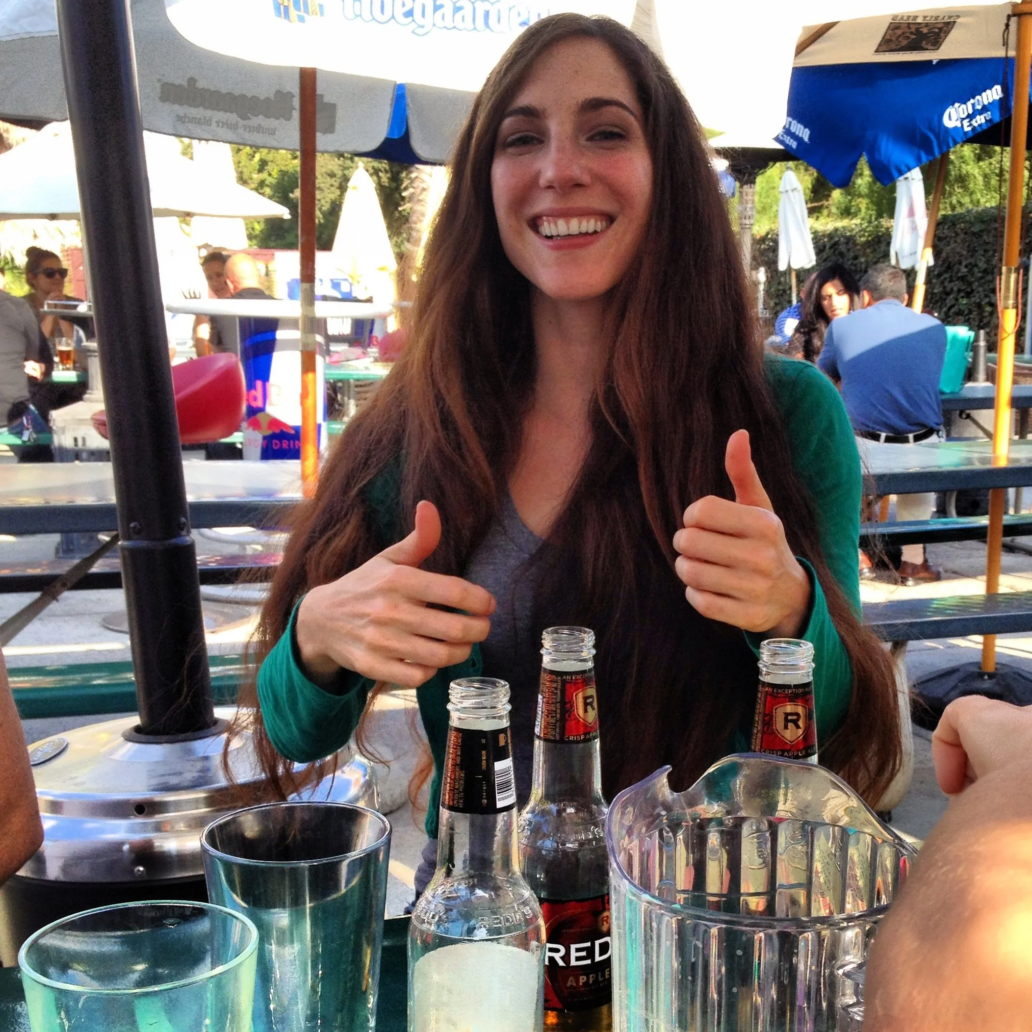 Brenda Norrie Padia with two thumbs up and a wide smile, wearing her long brown hair down and dark clothing. There are assorted bottles and pint glasses in the foreground.