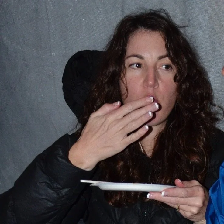 Michelle Brightstar snapped just after putting a large bite of delicious food in her mouth. One hand is covering her mouth. She is wearing her dark long hair down in a black puffer coat and is holding a plate.